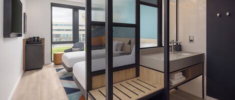 Quarto de hotel moderno com cama, escrivaninha, janela ampla, banheiro com pia e vista para o exterior.