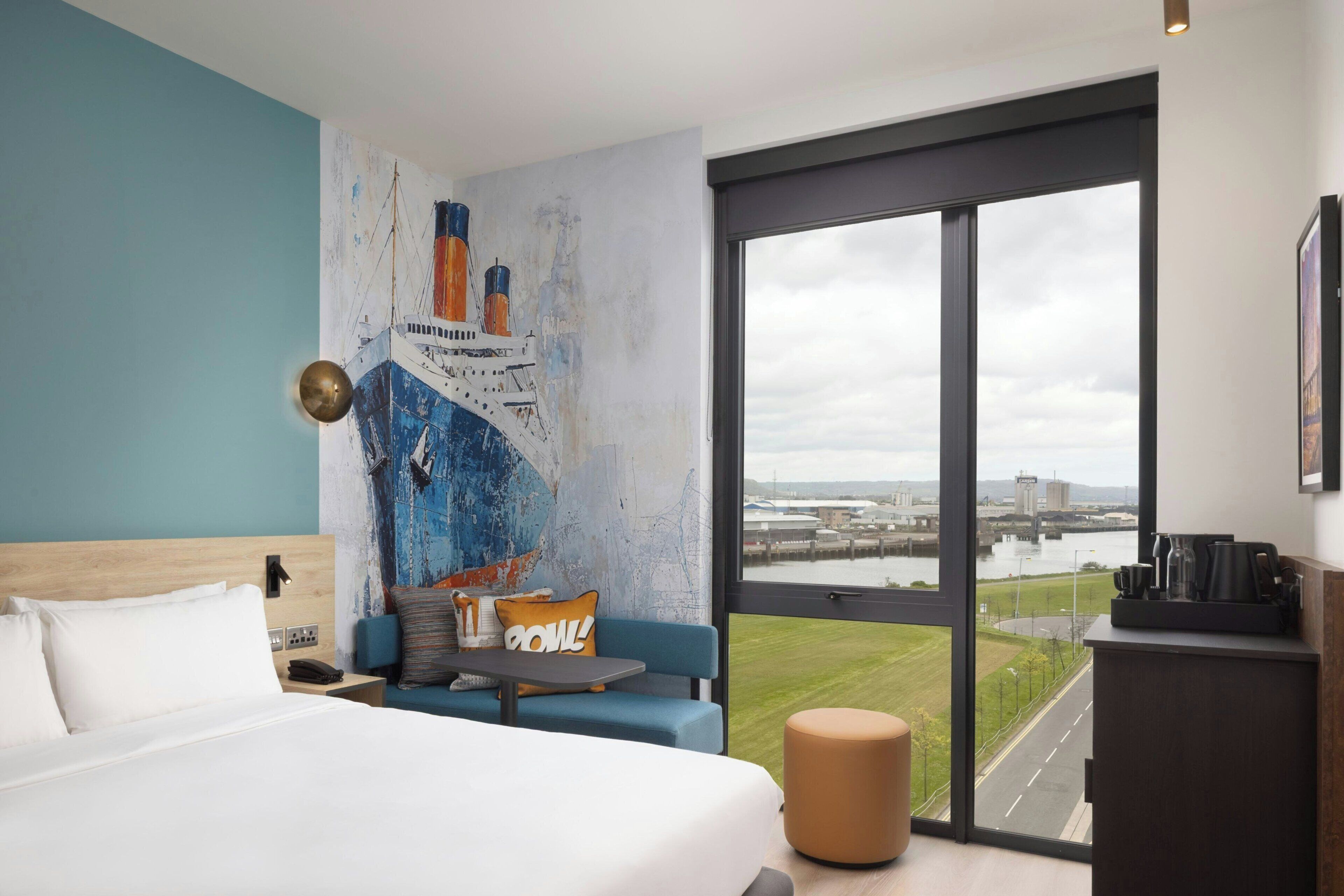 A modern hotel room with a large bed, a seating area with a view of the city, and a large window overlooking a river.