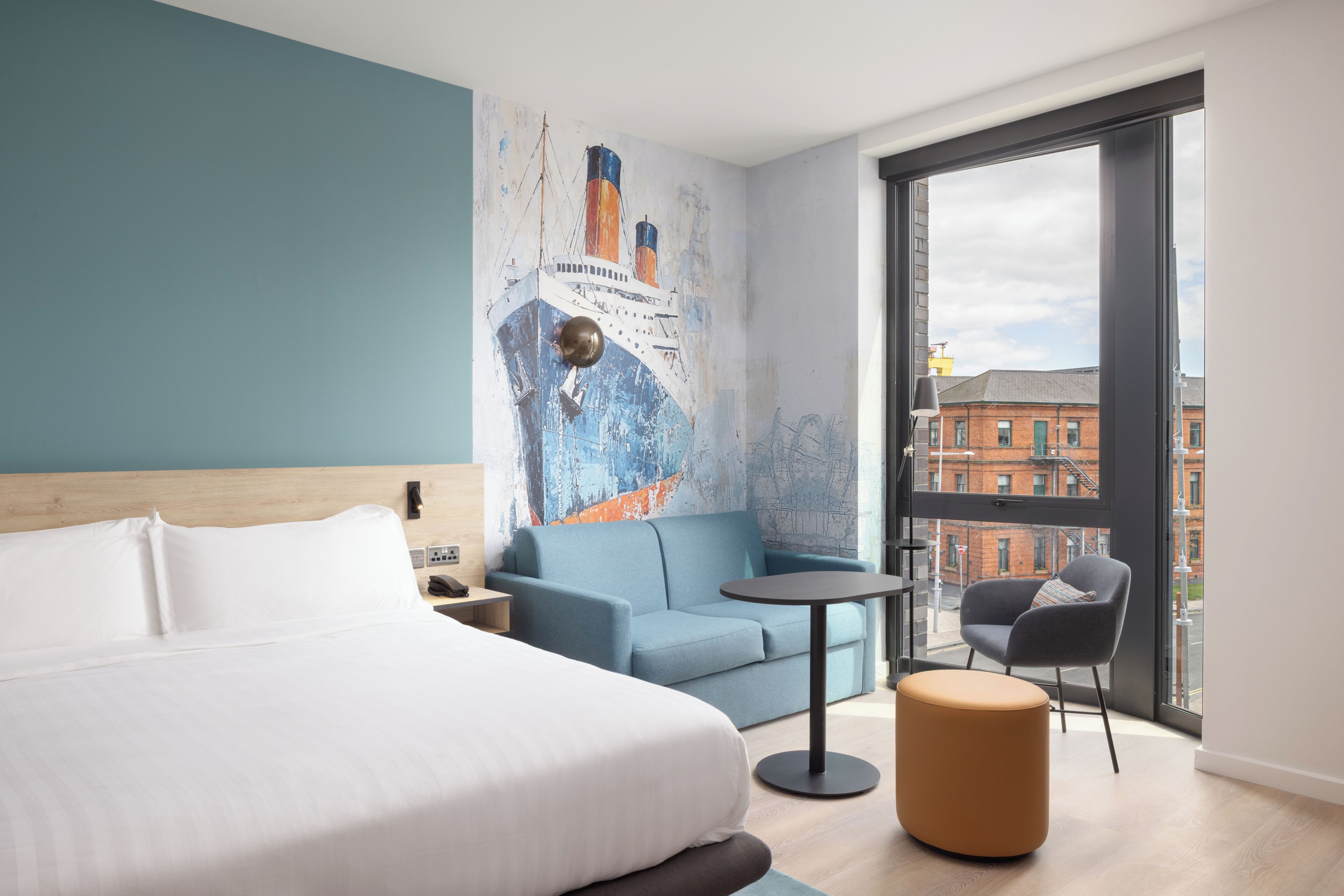 Una habitación de hotel moderna con una cama grande, un sofá azul, una mesita redonda y una silla.