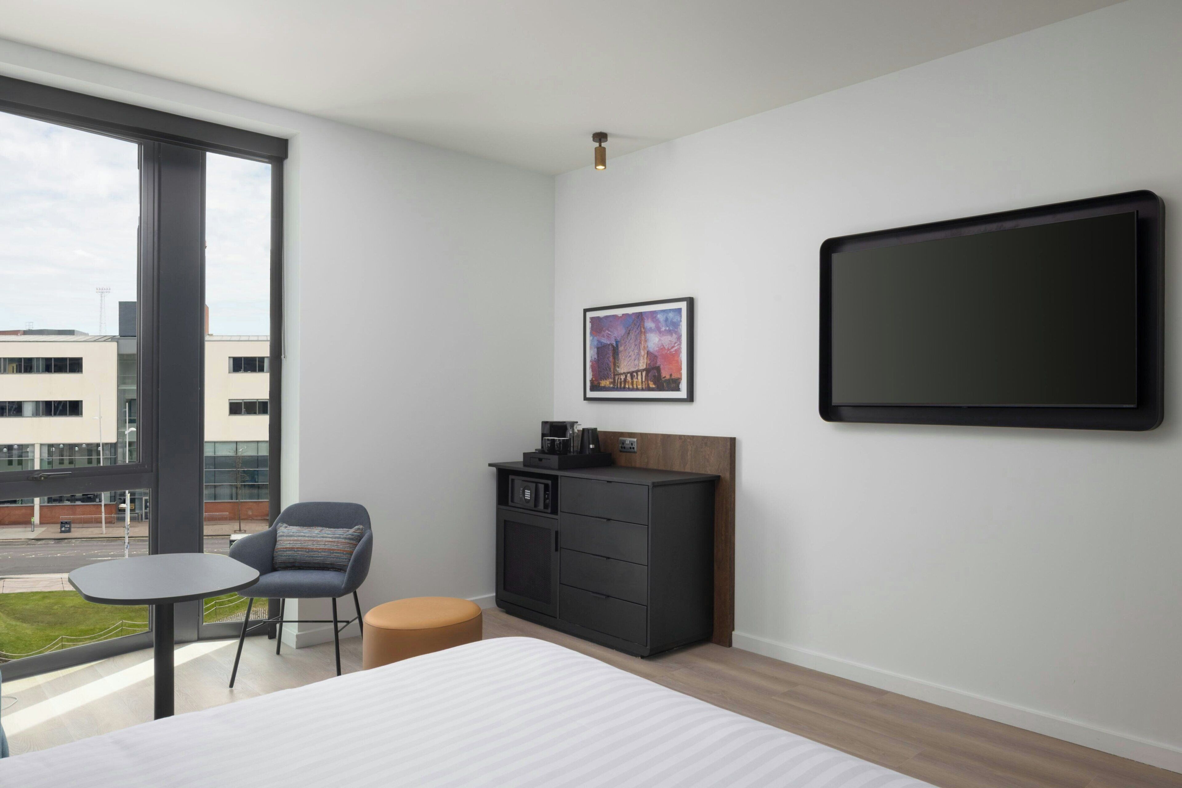 A modern hotel room with a large bed, a small table, a chair, a cabinet, a picture on the wall, and a large window.