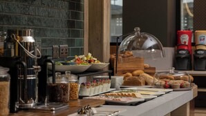 Un bufé con diversas opciones de comida, una estación de café y una vitrina de cristal con pasteles.