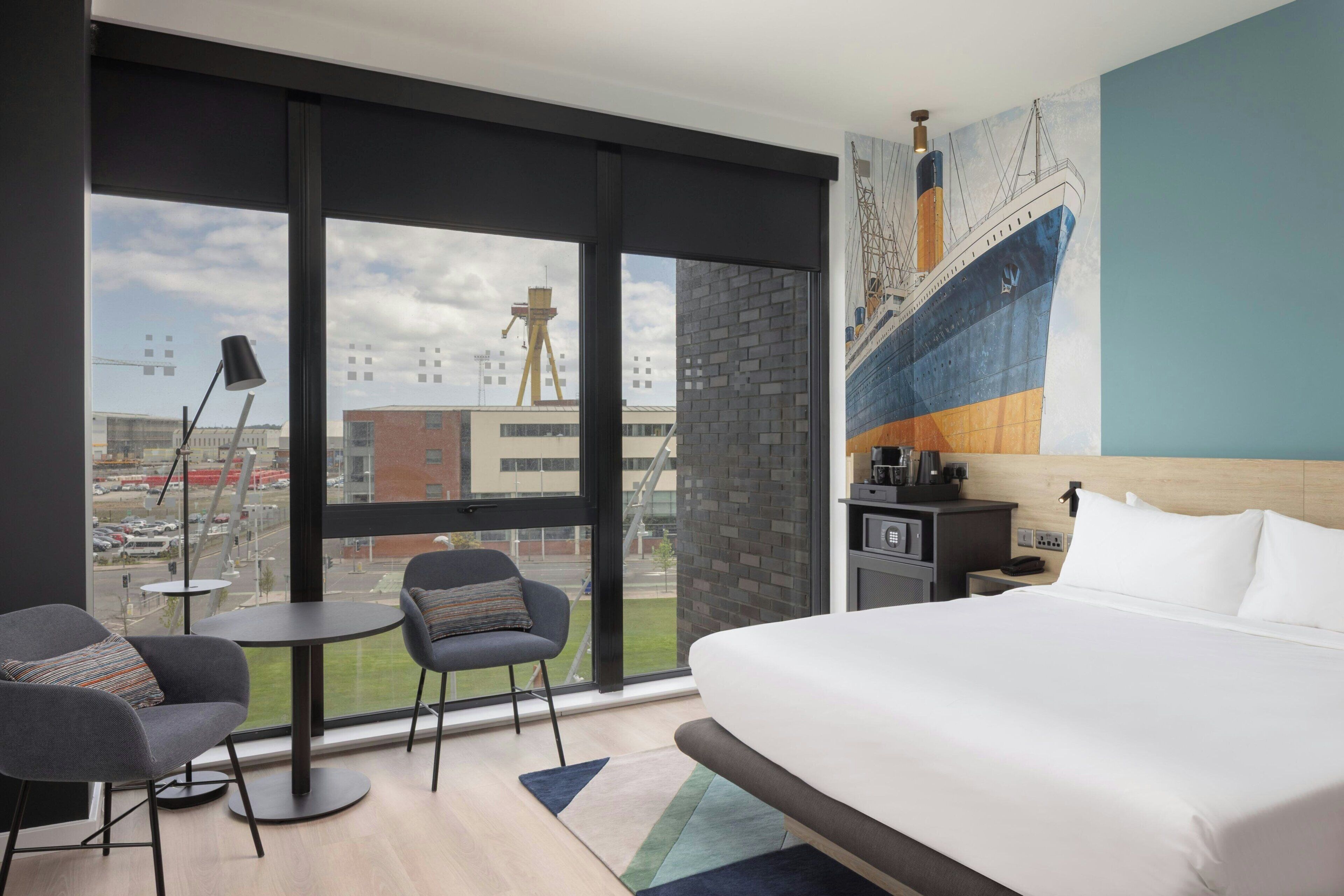 A modern hotel room with a large bed, two armchairs, a small table, a desk with a computer, and a large window with a city view.