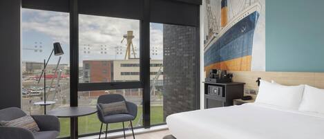 Moderni hotellihuone, jossa on suuri sänky, kaksi nojatuolia, pieni pöytä, työpöytä tietokoneella sekä suuri ikkuna, josta avautuu näkymä kaupunkiin.