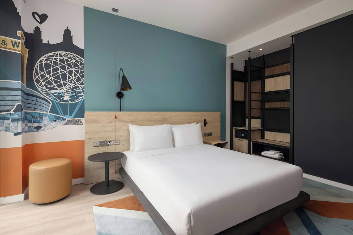 A modern hotel room with a large bed, a round side table, a wall mural, and built-in shelving.