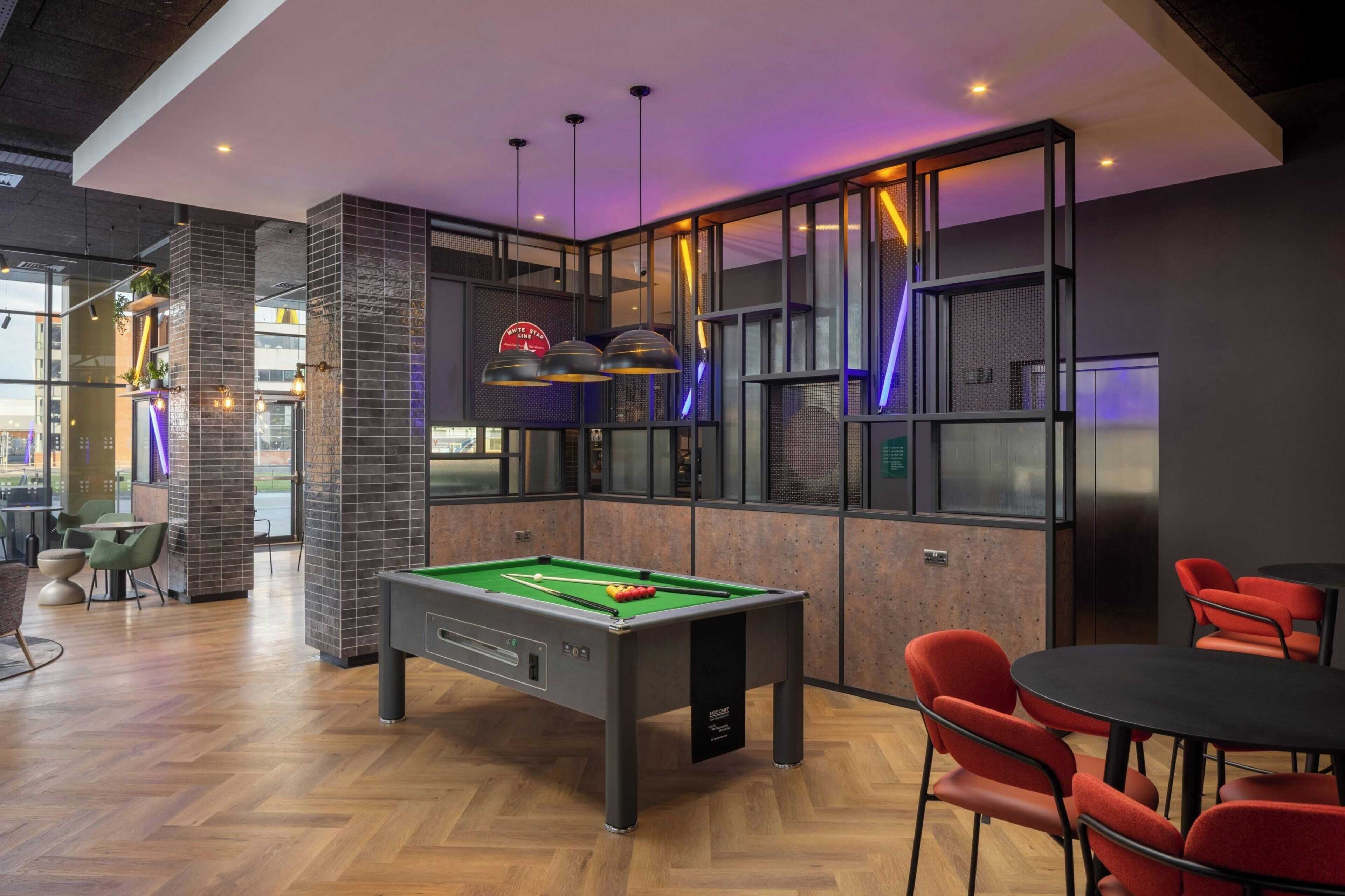 A pool table in a modern room with red chairs, a billiards rack, and a bar area with stools.