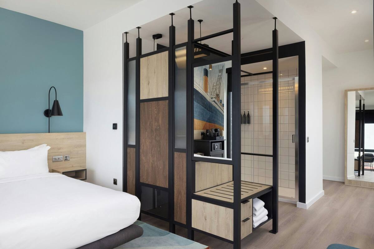 A modern hotel room with a bed, a wooden headboard, a partition with a view of a kitchen, and a bathroom with a mirror.