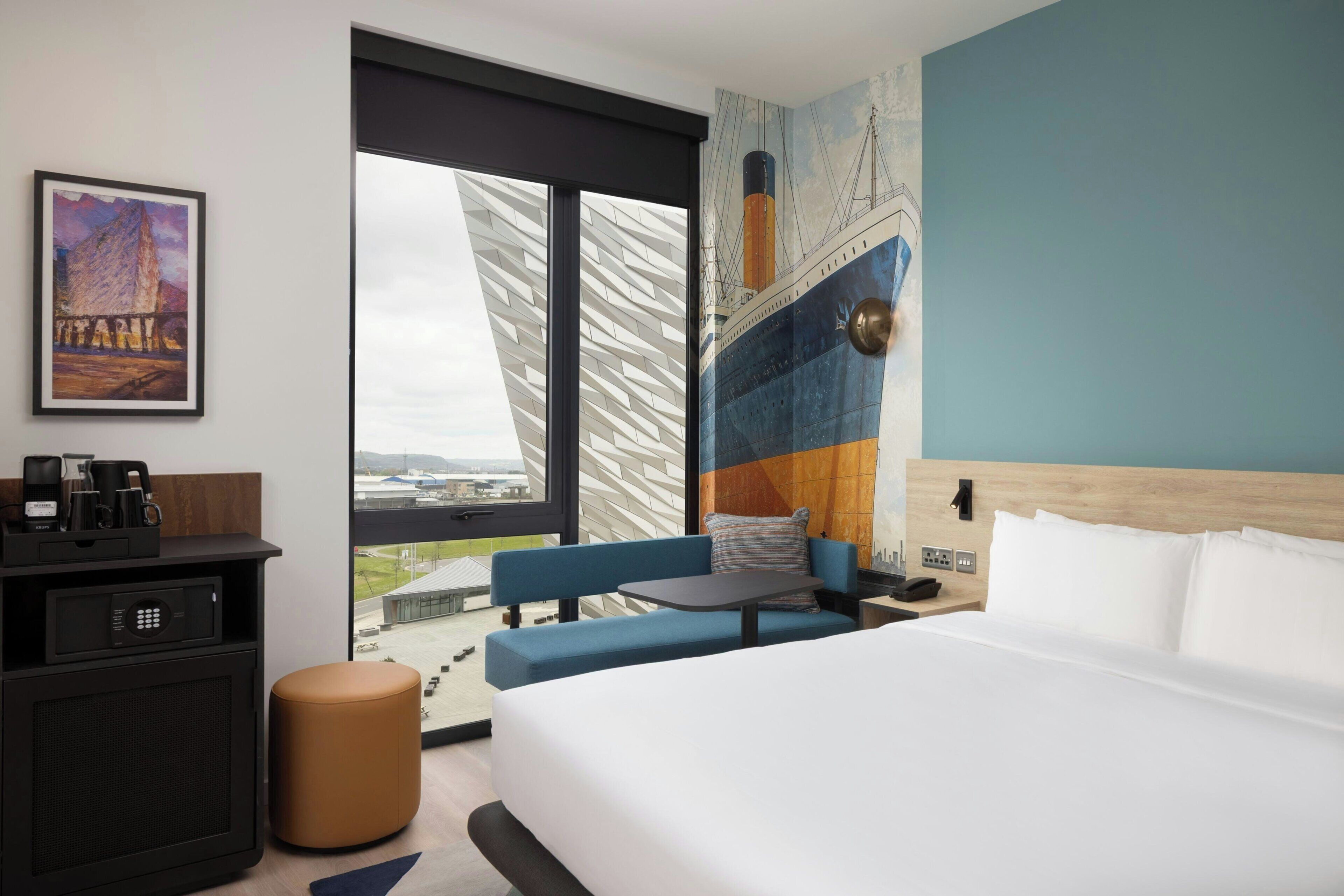 A hotel room with a bed, a sofa, a small table, a coffee maker, a TV, and a large window with a view.