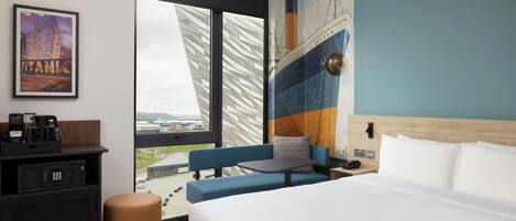 A hotel room with a bed, a sofa, a small table, a coffee maker, a TV, and a large window with a view.