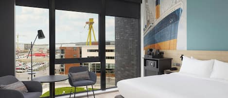 Une chambre d’hôtel avec un grand lit, deux fauteuils, une petite table, un bureau avec une lampe et une fresque représentant un navire sur le mur.