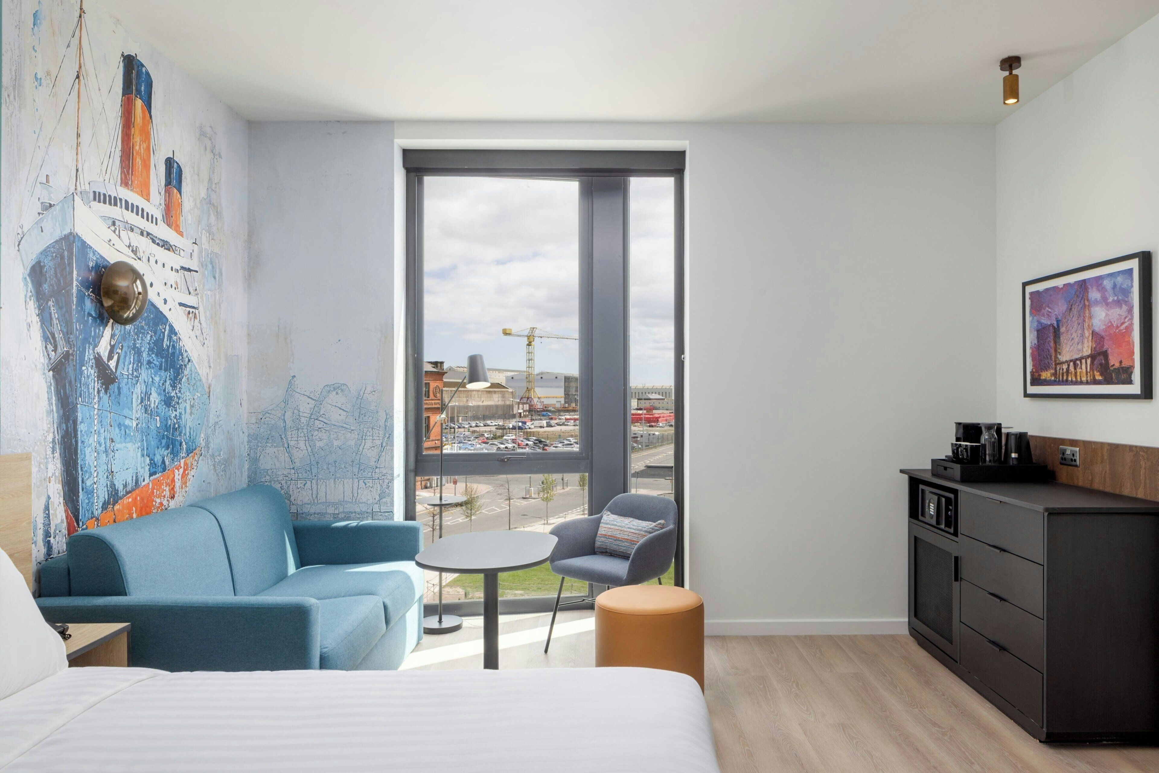 A modern hotel room with a large window, a sofa, a small table, a chair, a bed, and a minibar.