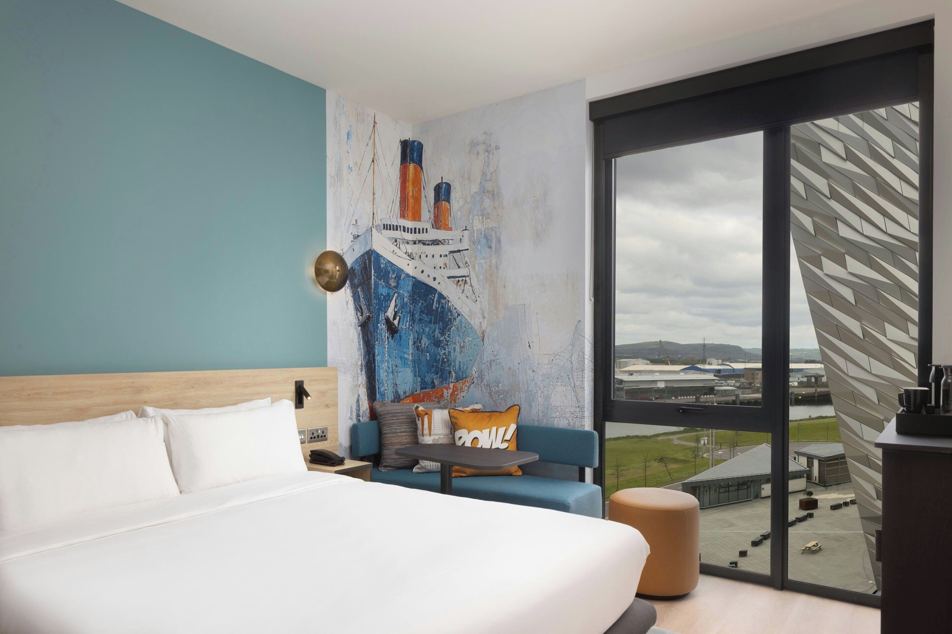 A hotel room with a large bed, a blue sofa, a small table, and a view of an airport through a large window.