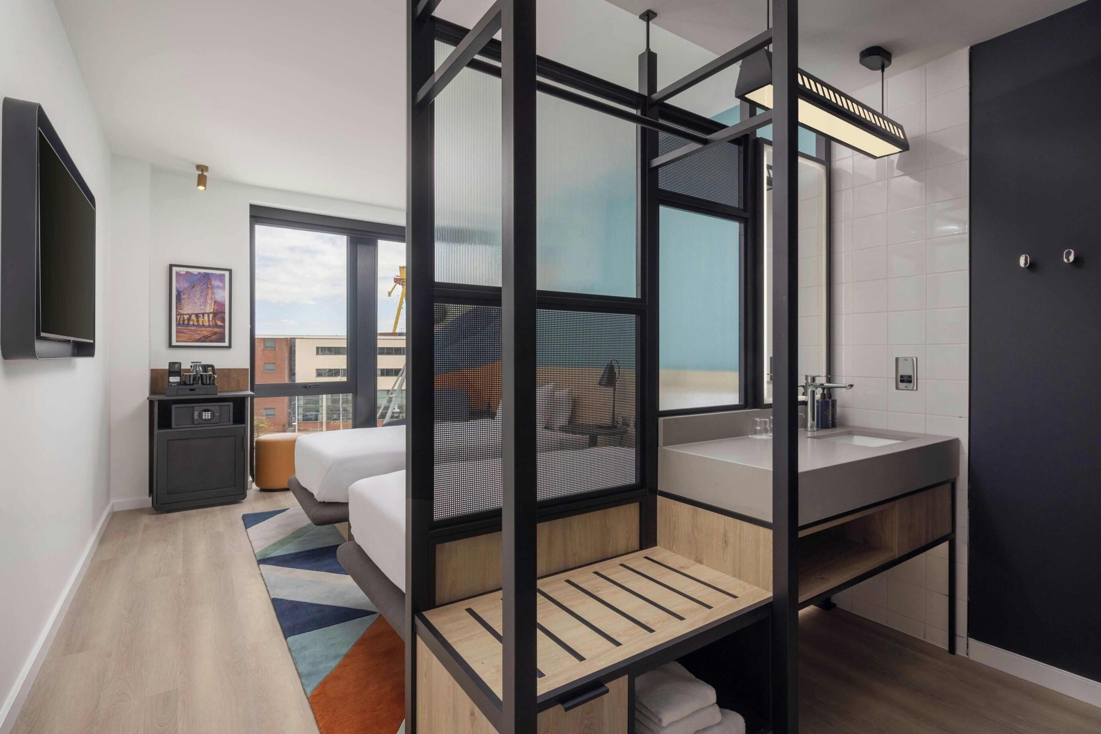 A modern hotel room with a bed, a desk, a large window, and a bathroom.