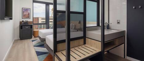 A modern hotel room with a bed, a desk, a large window, and a bathroom.