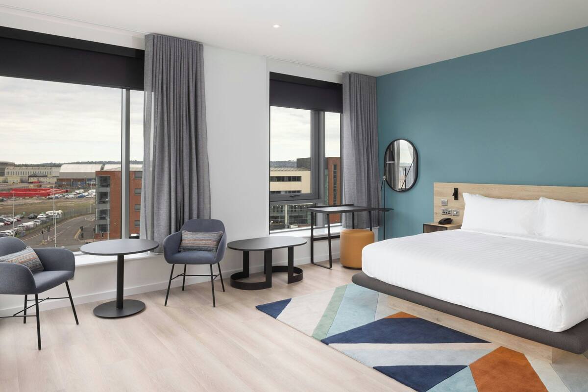 A modern hotel room with a large bed, two chairs, a small table, and a view of the cityscape.