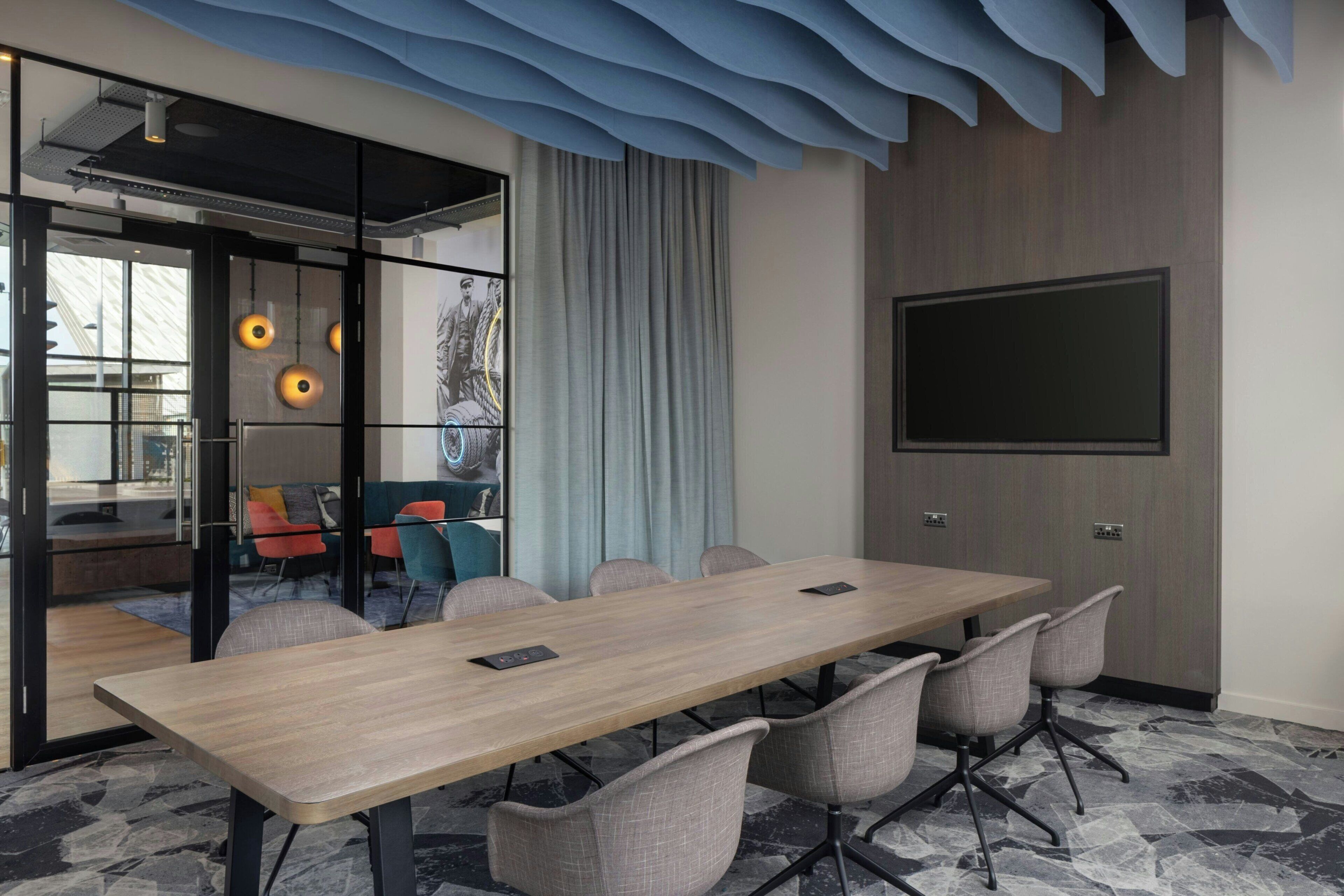 A conference room with a long table, six chairs, a large flat-screen TV, and a glass partition with a view.