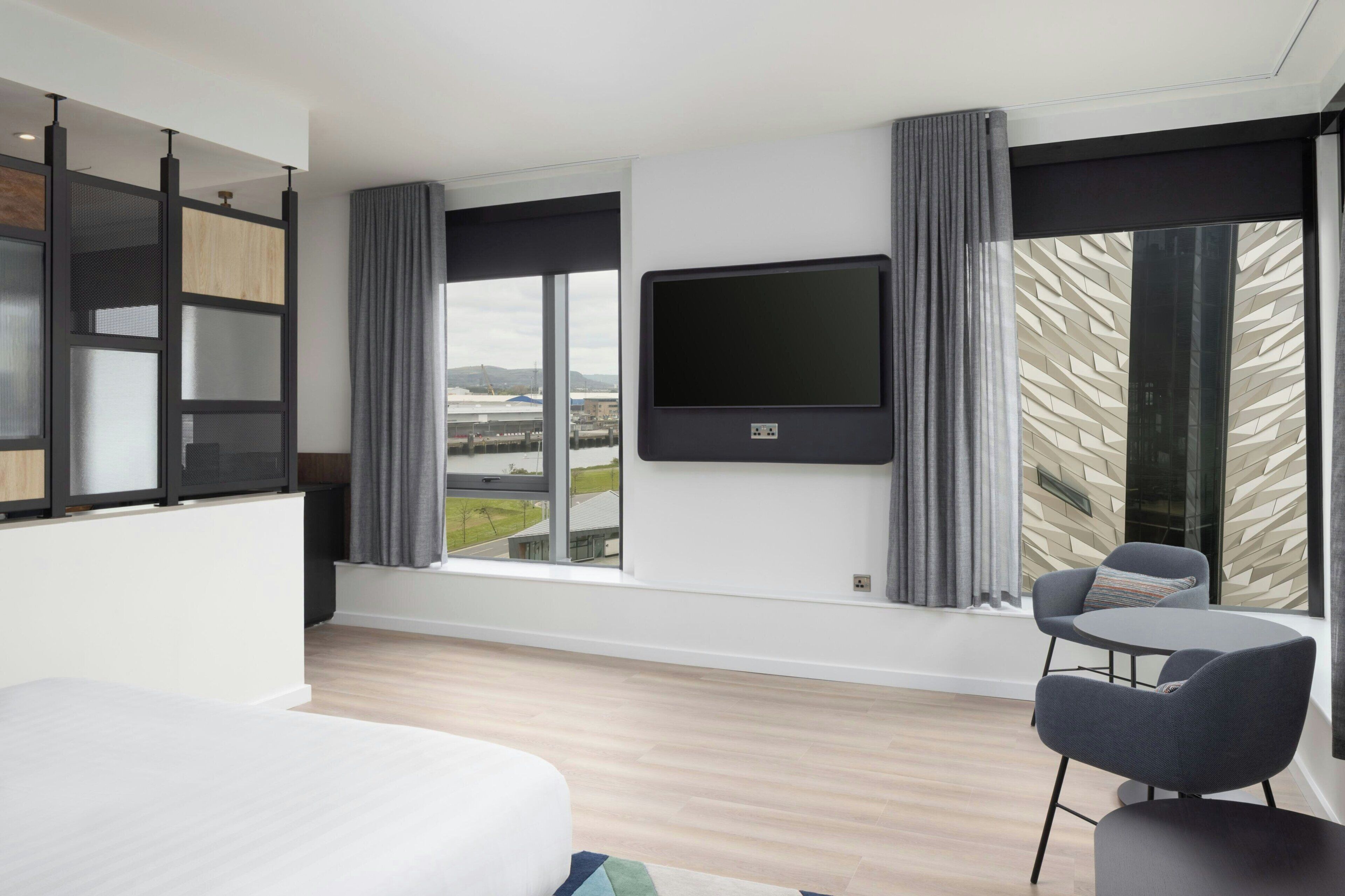 A modern hotel room with a large bed, a TV mounted on the wall, a seating area with two chairs, and a large window offering a view of the cityscape.