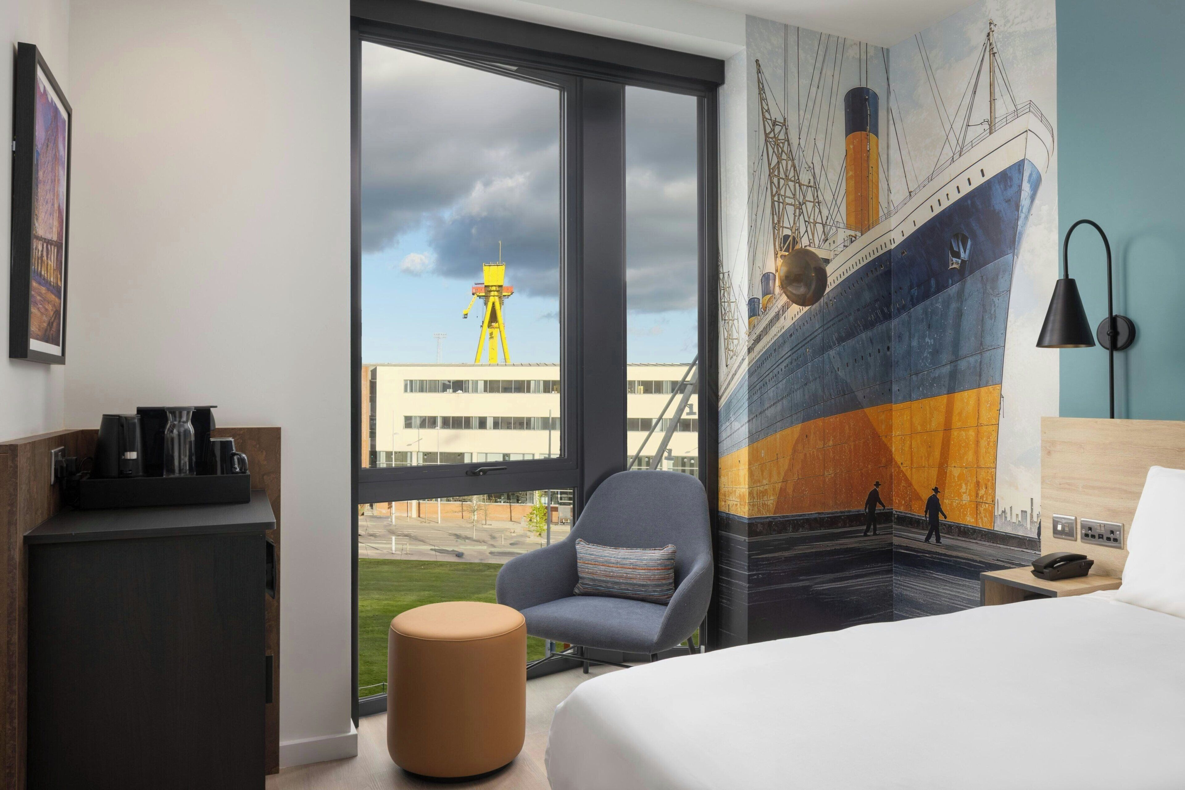A hotel room with a large window, a bed, a chair, a small ottoman, a desk with a phone, and a mural of a ship.