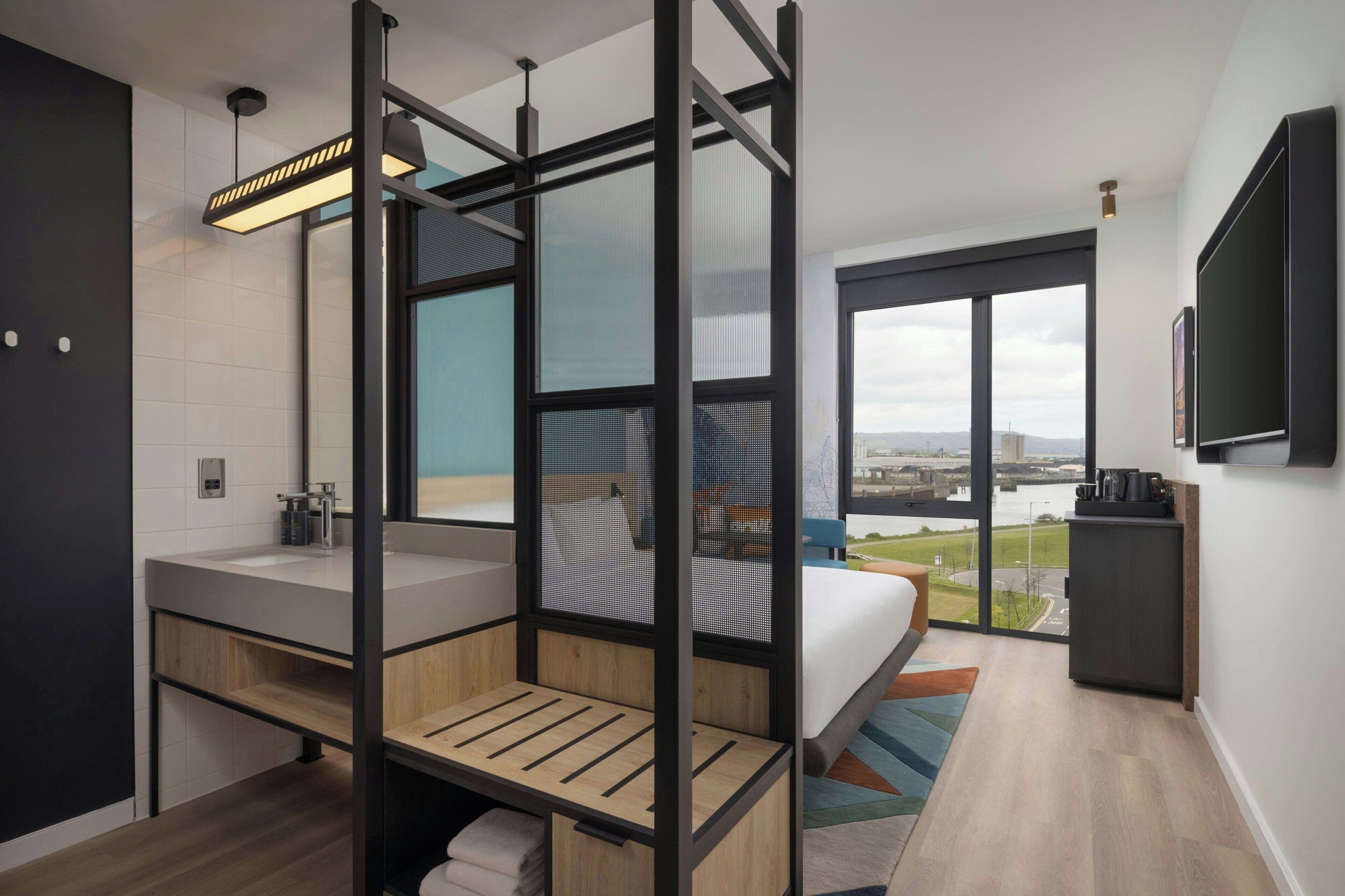 Una habitación de hotel moderna con un ventanal grande, una cama, un baño con lavabo y un televisor montado en la pared.