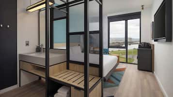 Una habitación de hotel moderna con un ventanal grande, una cama, un baño con lavabo y un televisor montado en la pared.