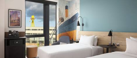 Habitación de hotel con dos camas, un escritorio con cafetera, una silla, una mesita de noche con lámpara y un ventanal con vista a un barco y una grúa.