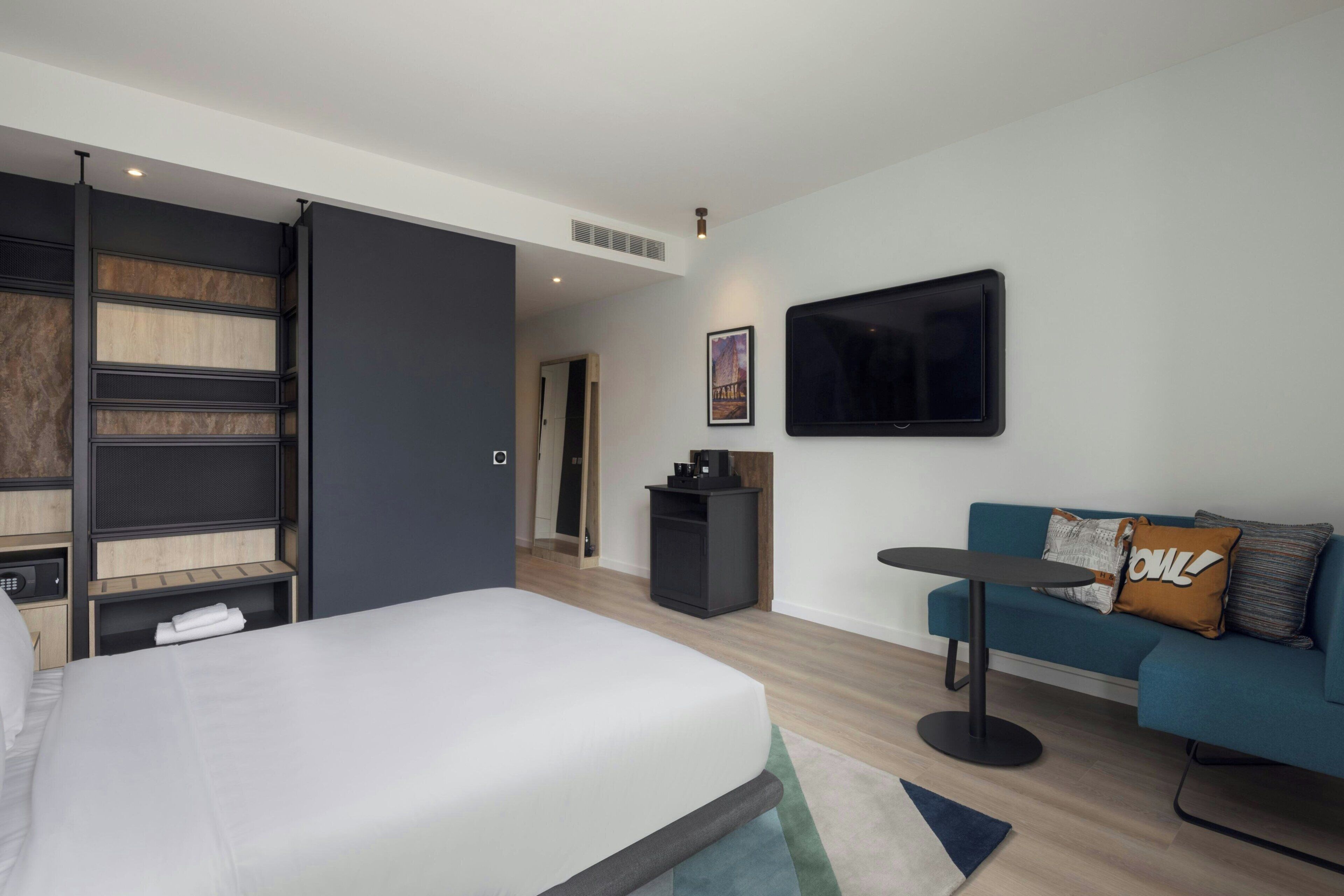 A modern hotel room with a bed, a sofa, a small table, a TV, and a desk with a computer.