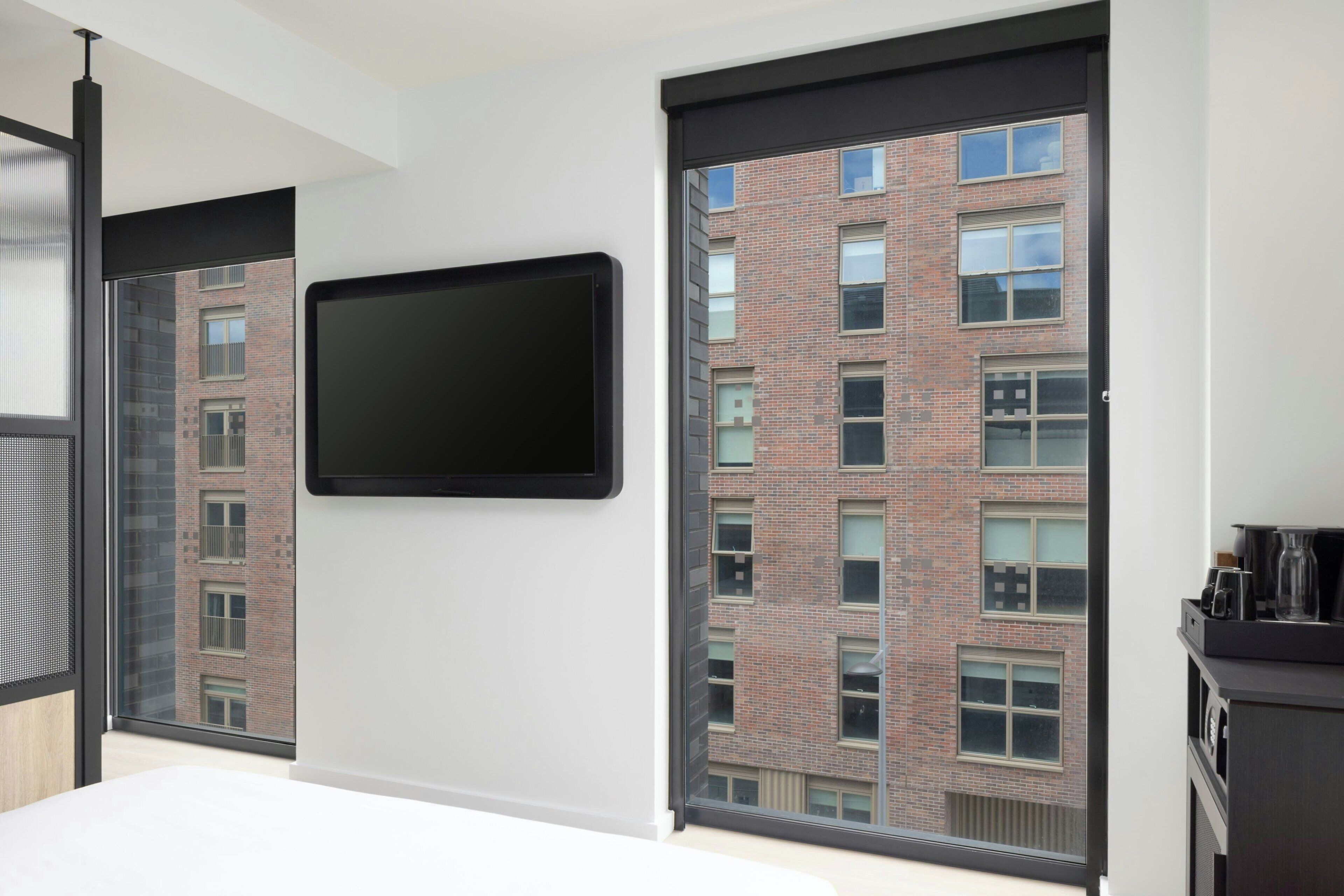 A modern hotel room with a large window, a flat-screen TV mounted on the wall, and a view of a brick building outside.