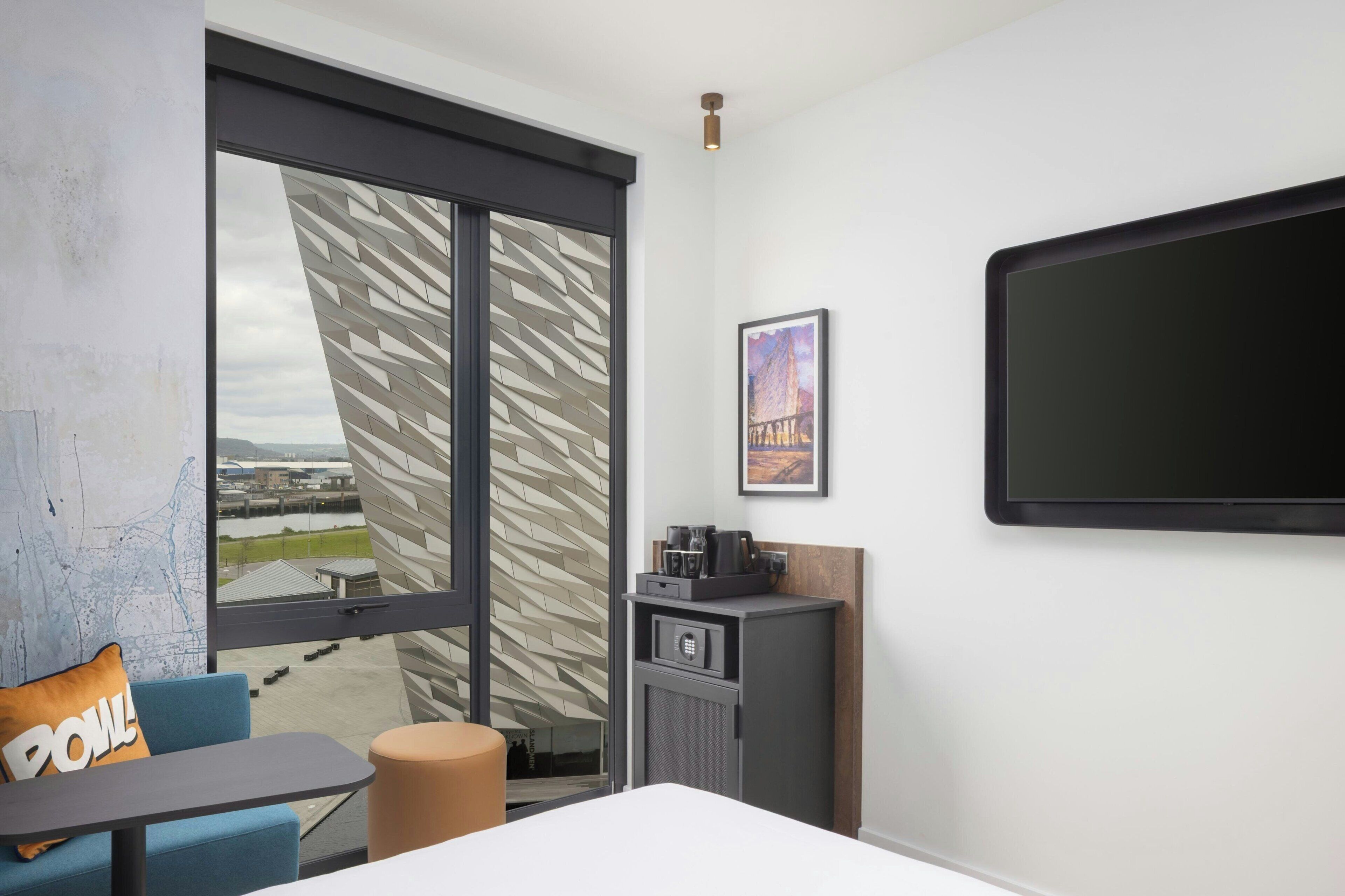 A hotel room with a large window, a flat-screen TV, a small cabinet, a sofa, and a table.