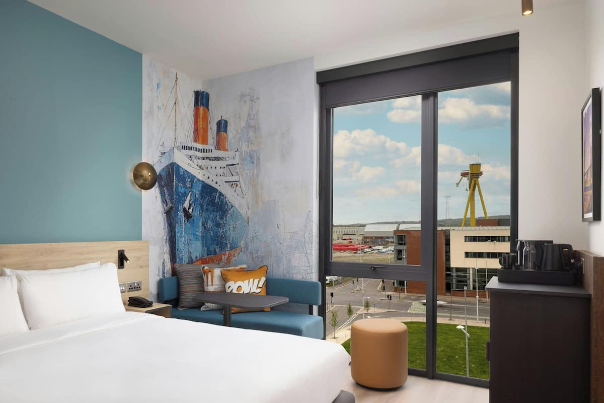 A modern hotel room with a large bed, a seating area with a view, and a mural of a ship on the wall.