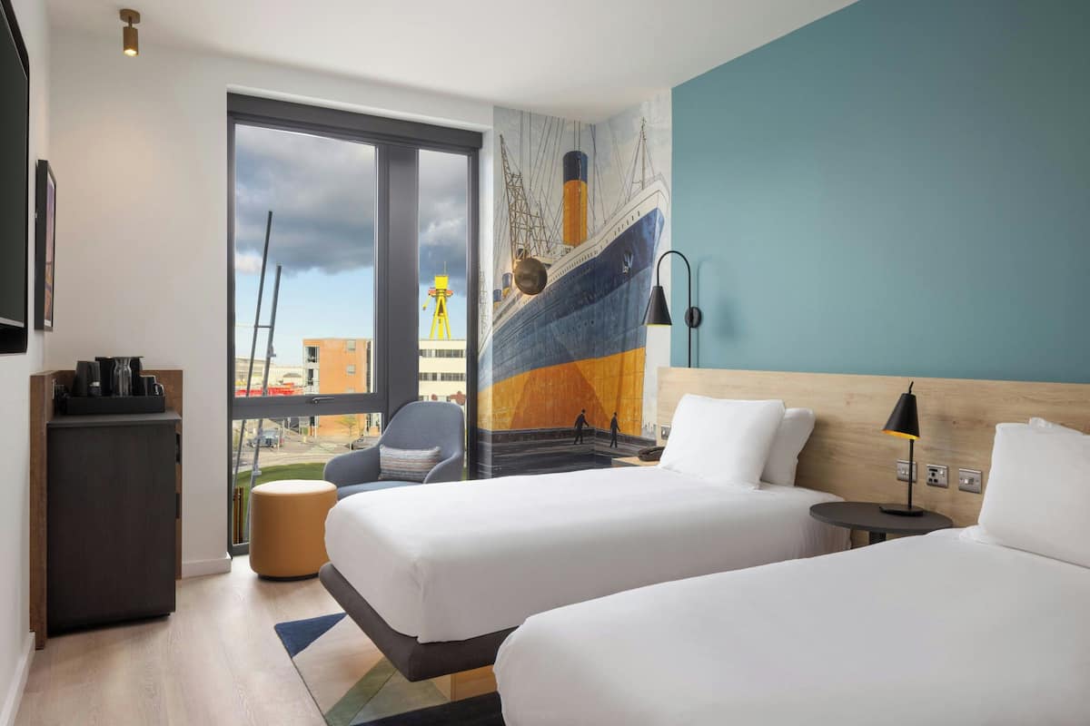 A hotel room with two beds, a desk, a chair, a small table with a lamp, and a large window with a view of the outside.