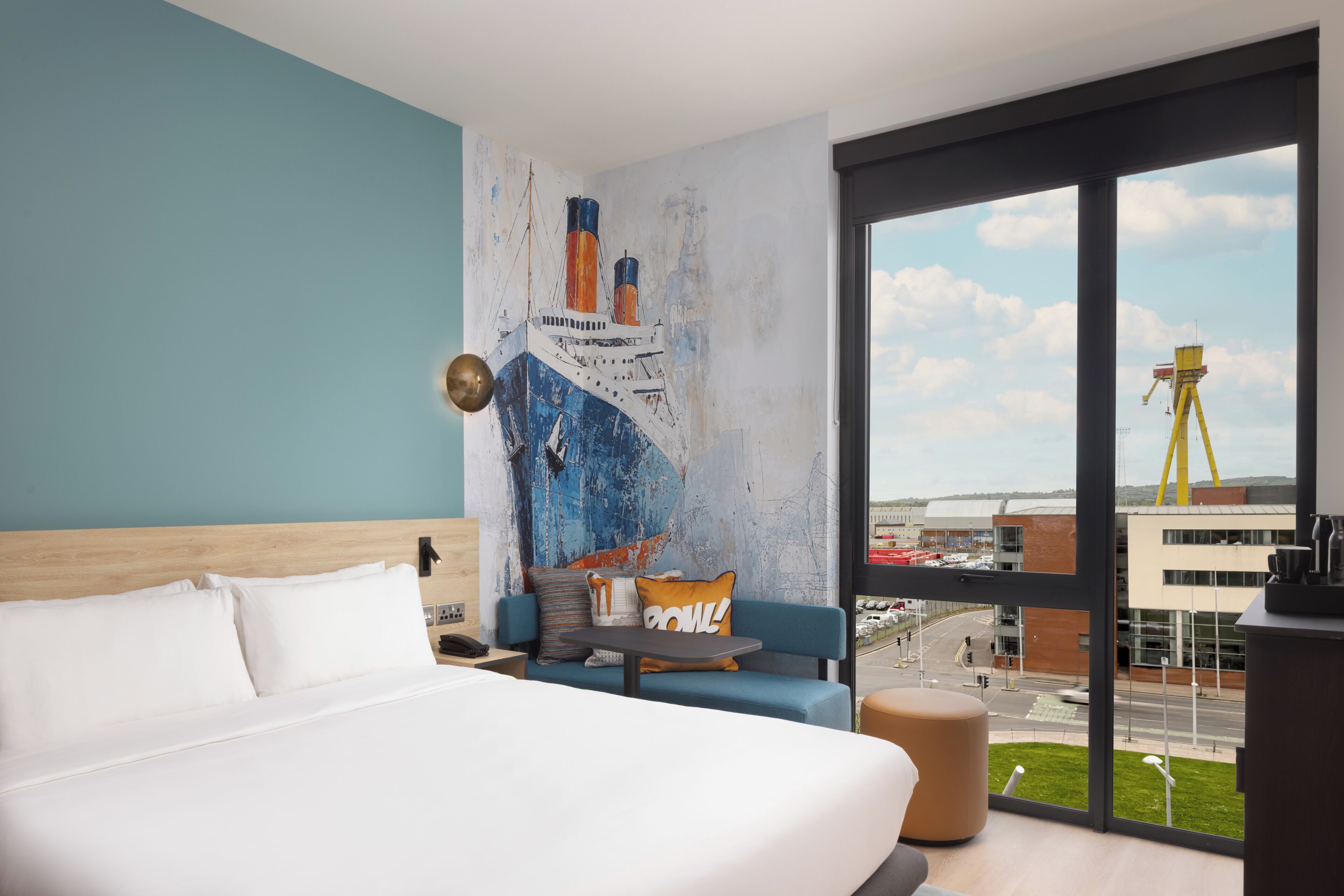 Une chambre d’hôtel avec un grand lit, un canapé, une petite table et une vue sur le paysage urbain, grâce à de grandes fenêtres.