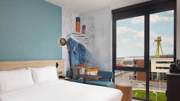 Habitación de hotel con una cama grande, un sofá, una mesita y vistas a un paisaje urbano a través de amplias ventanas.