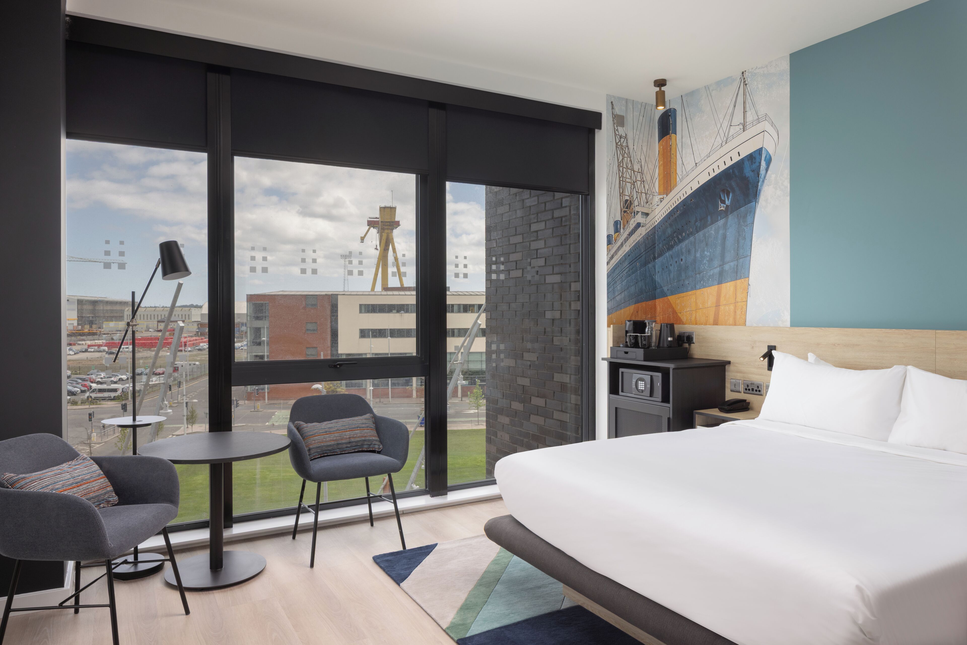 A hotel room with a bed, two chairs, a small table, a lamp, a painting, and a view of the city.