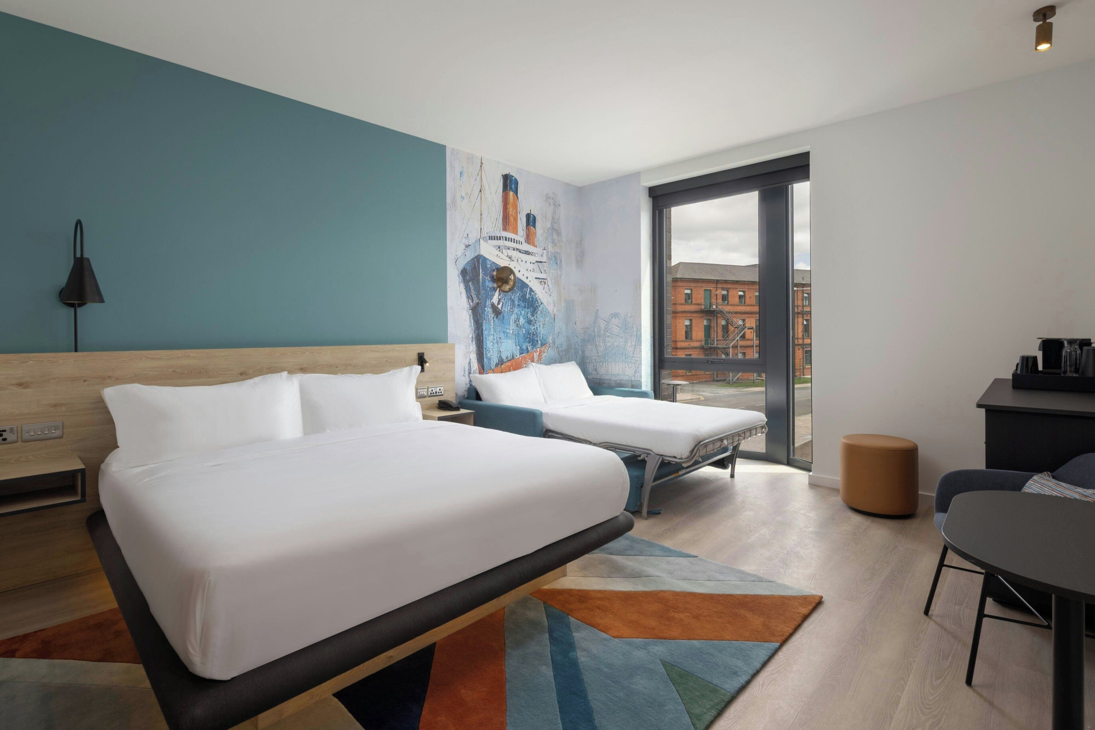 A hotel room with two beds, a desk, a chair, a lamp, and a large window with a view of buildings.