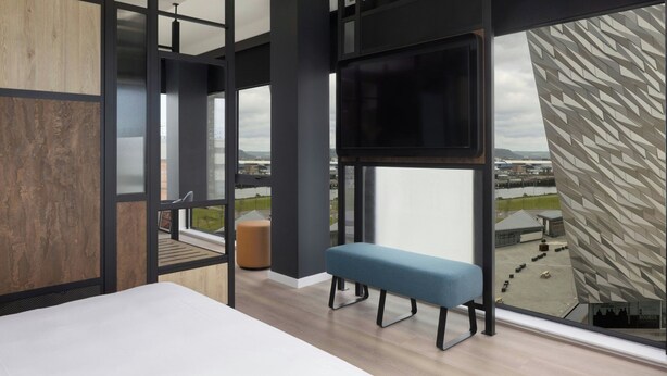 Una habitación de hotel moderna con un ventanal grande, un televisor de pantalla plana, un banco y vistas a la ciudad.