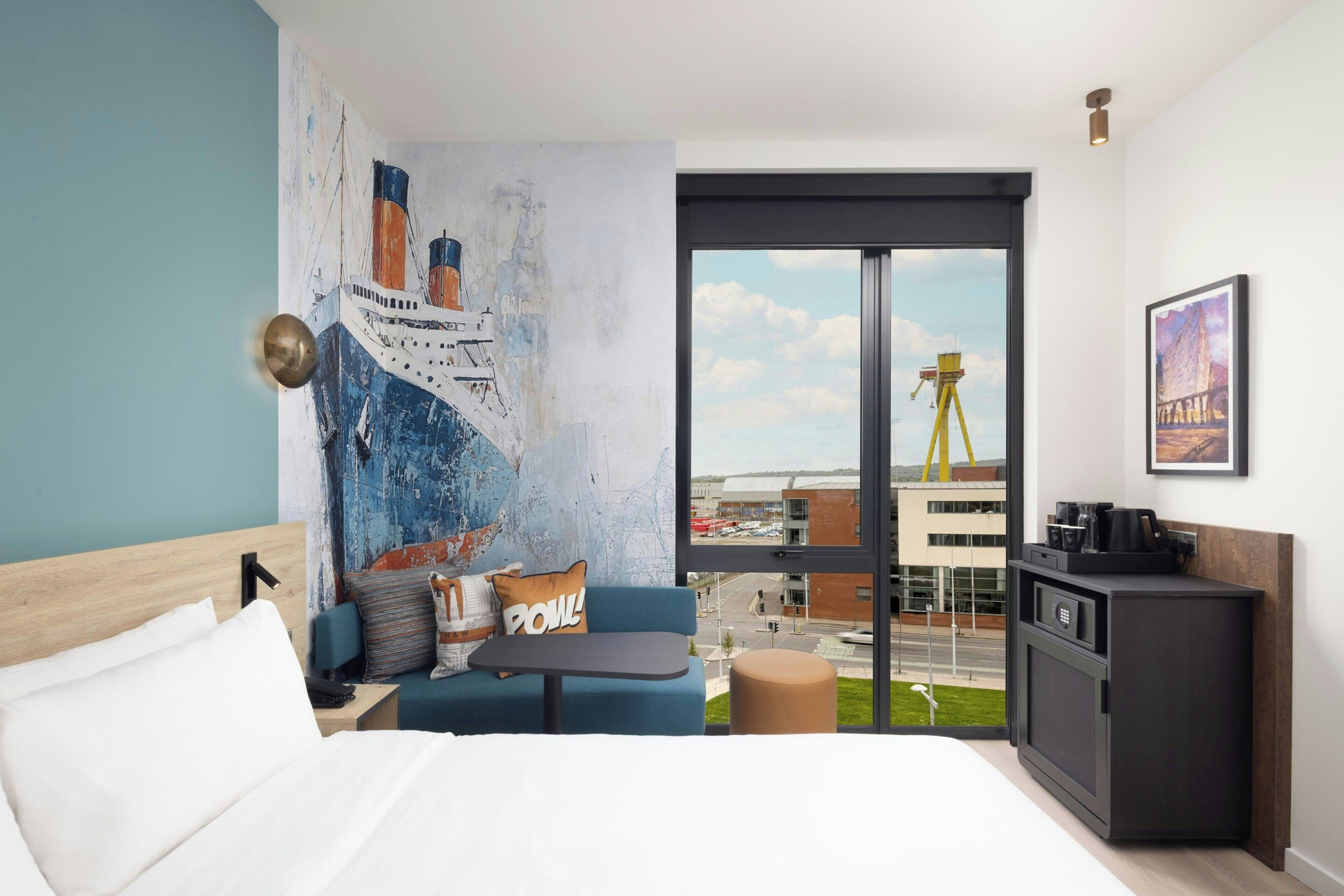 Una habitación de hotel moderna con una cama grande, una mesita pequeña, un sofá, un escritorio, un armario y un ventanal con vistas a un paisaje urbano y una grúa.