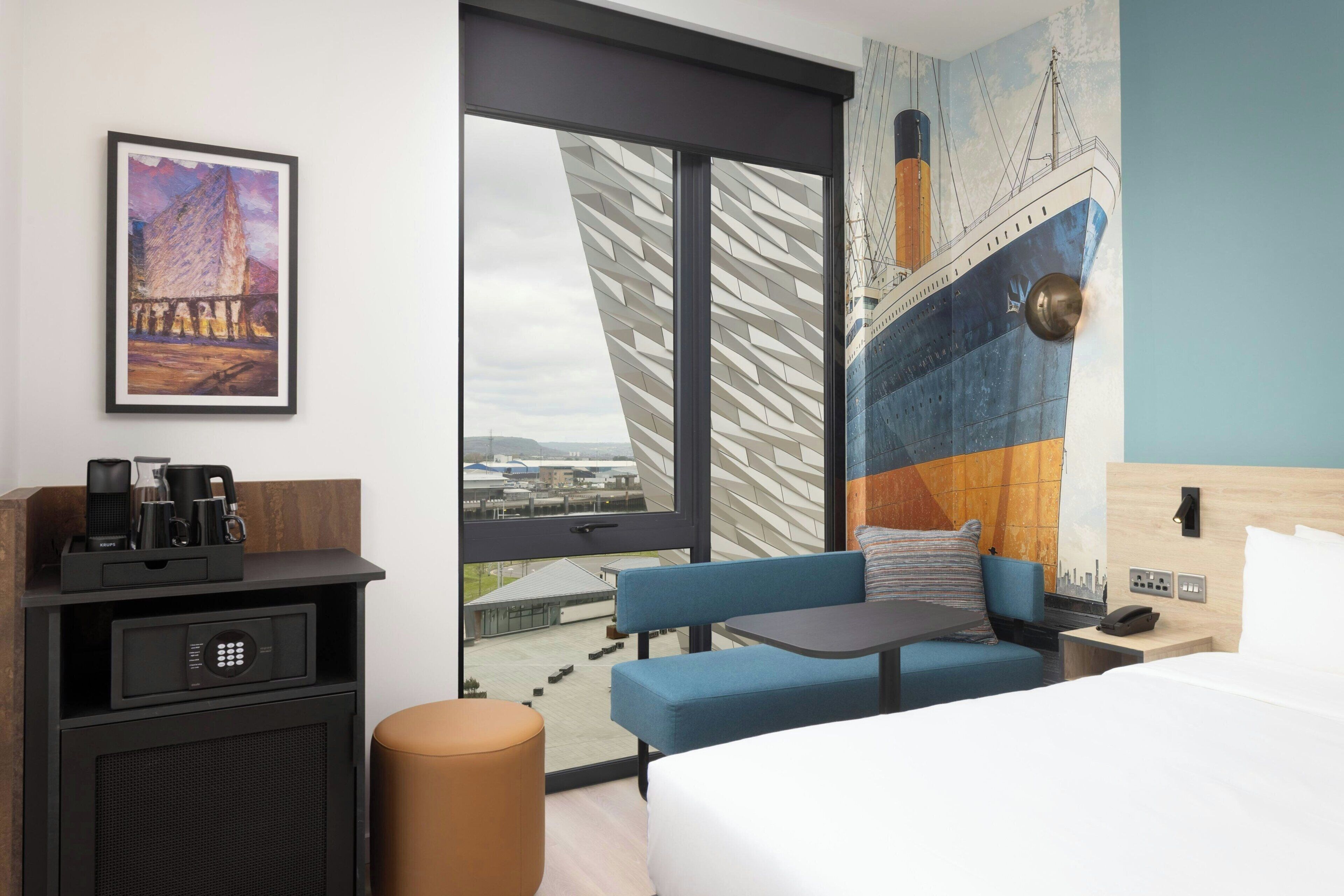 A hotel room with a bed, a desk, a chair, a sofa, a coffee maker, a phone, and a view of a cityscape.