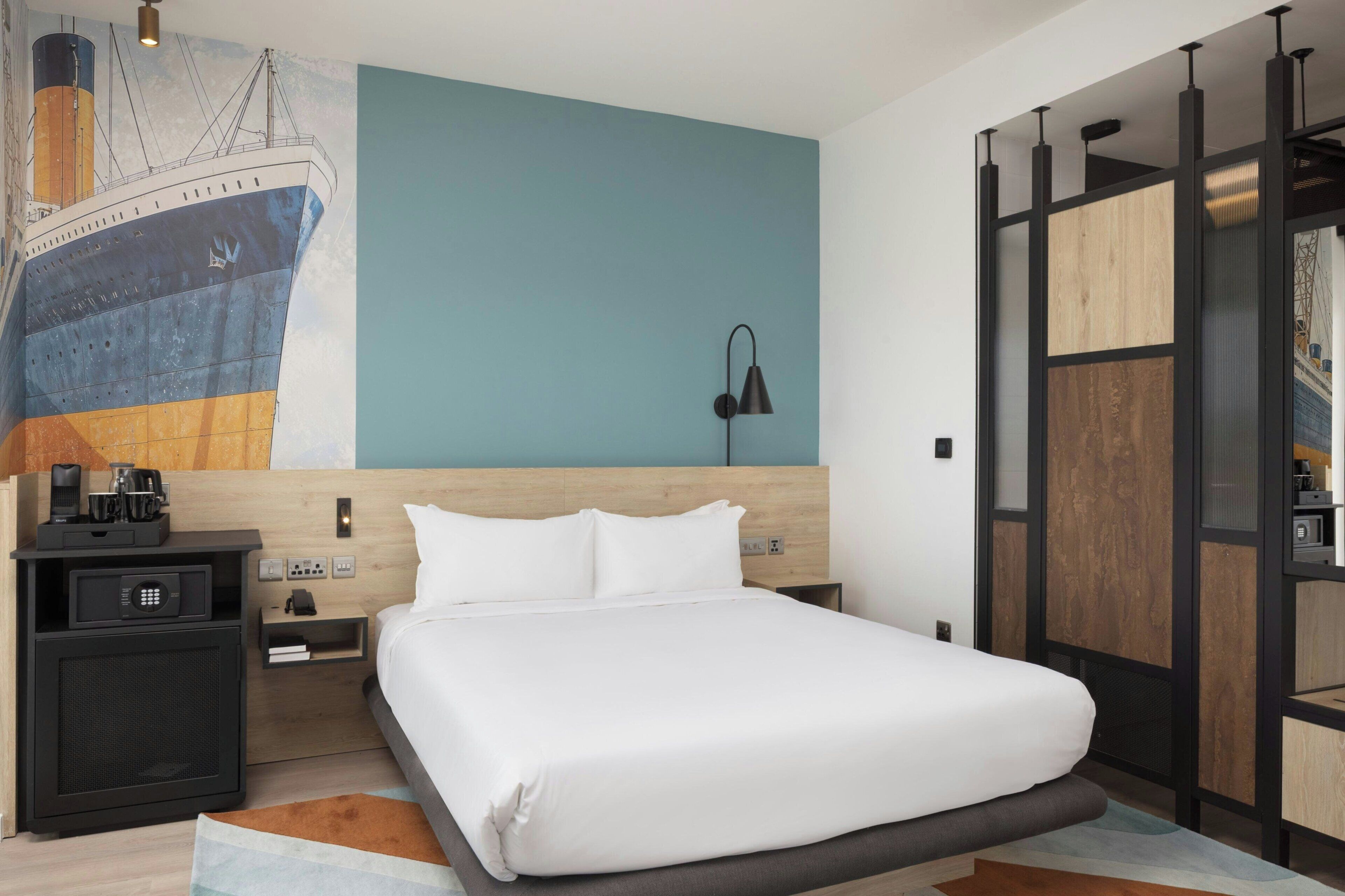 Habitación de hotel moderna con una cama grande, una mesita de noche, una lámpara y un mural en la pared con un barco.