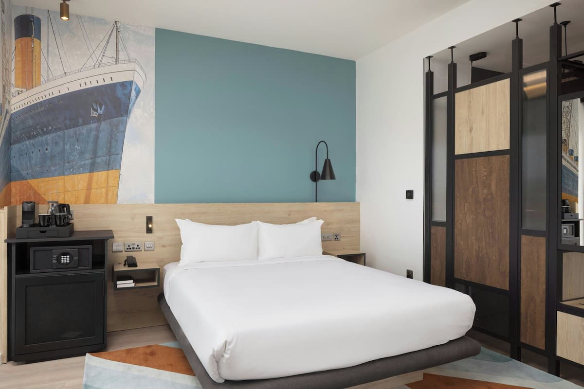 A modern hotel room with a large bed, a nightstand, a lamp, and a wall mural of a ship.