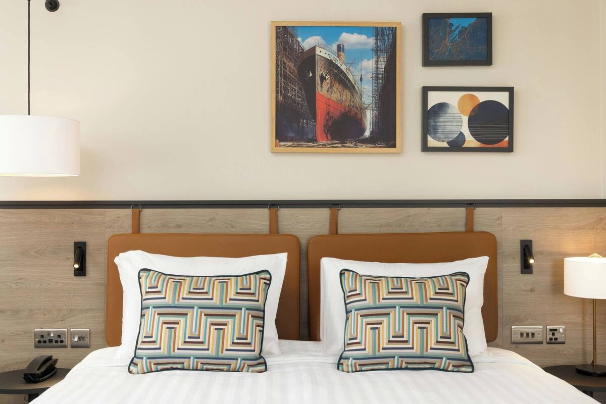 A hotel room with a bed, two patterned pillows, a nightstand, a phone, a lamp, and three framed artworks on the wall.