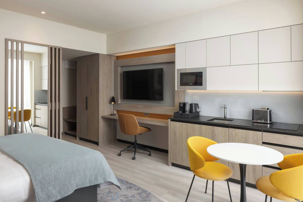 A modern hotel room with a bed, a desk, a TV, a microwave, and a dining area with yellow chairs.