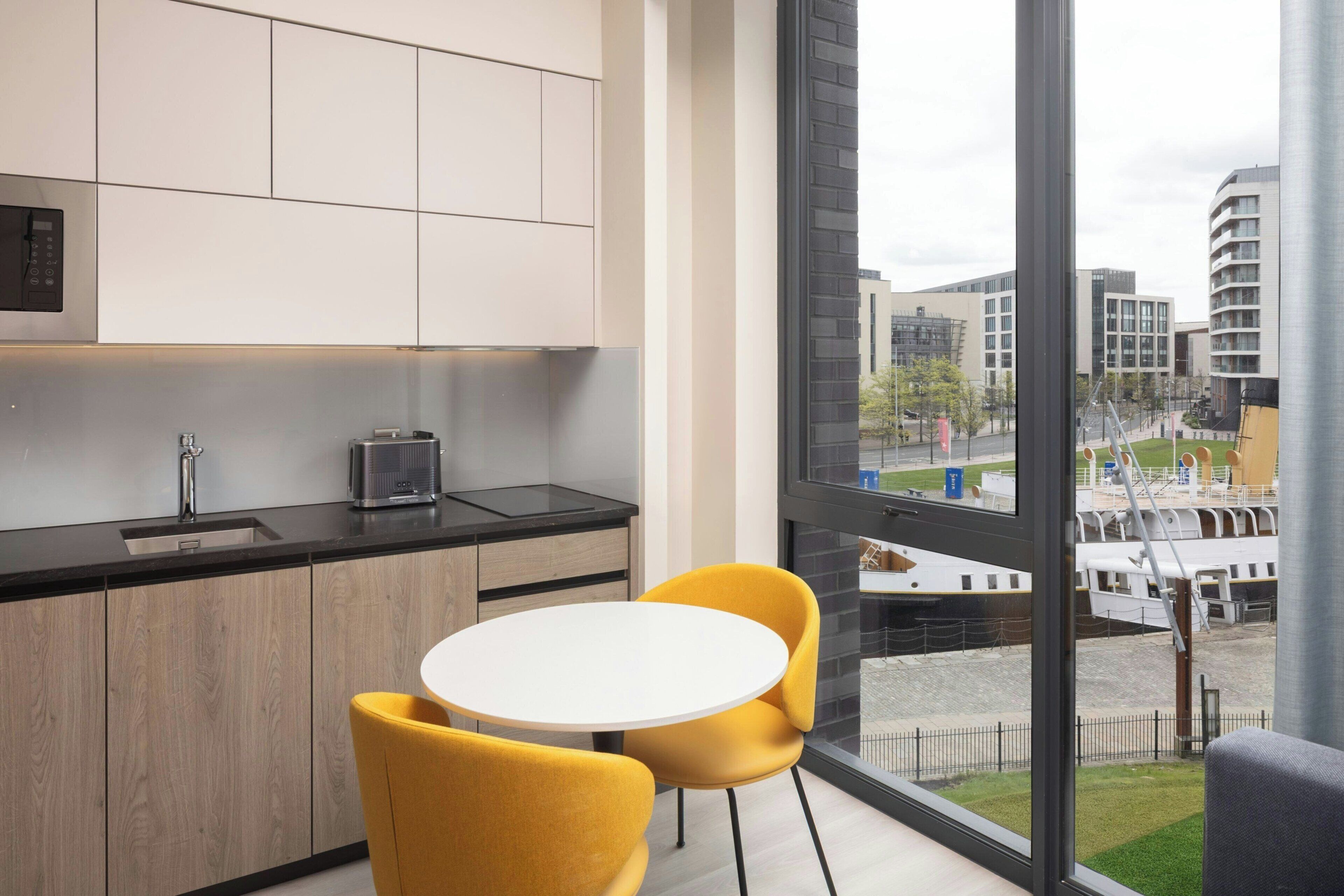 Una cocina moderna con un amplio ventanal, una mesa redonda con sillas amarillas y vistas a un paisaje urbano.