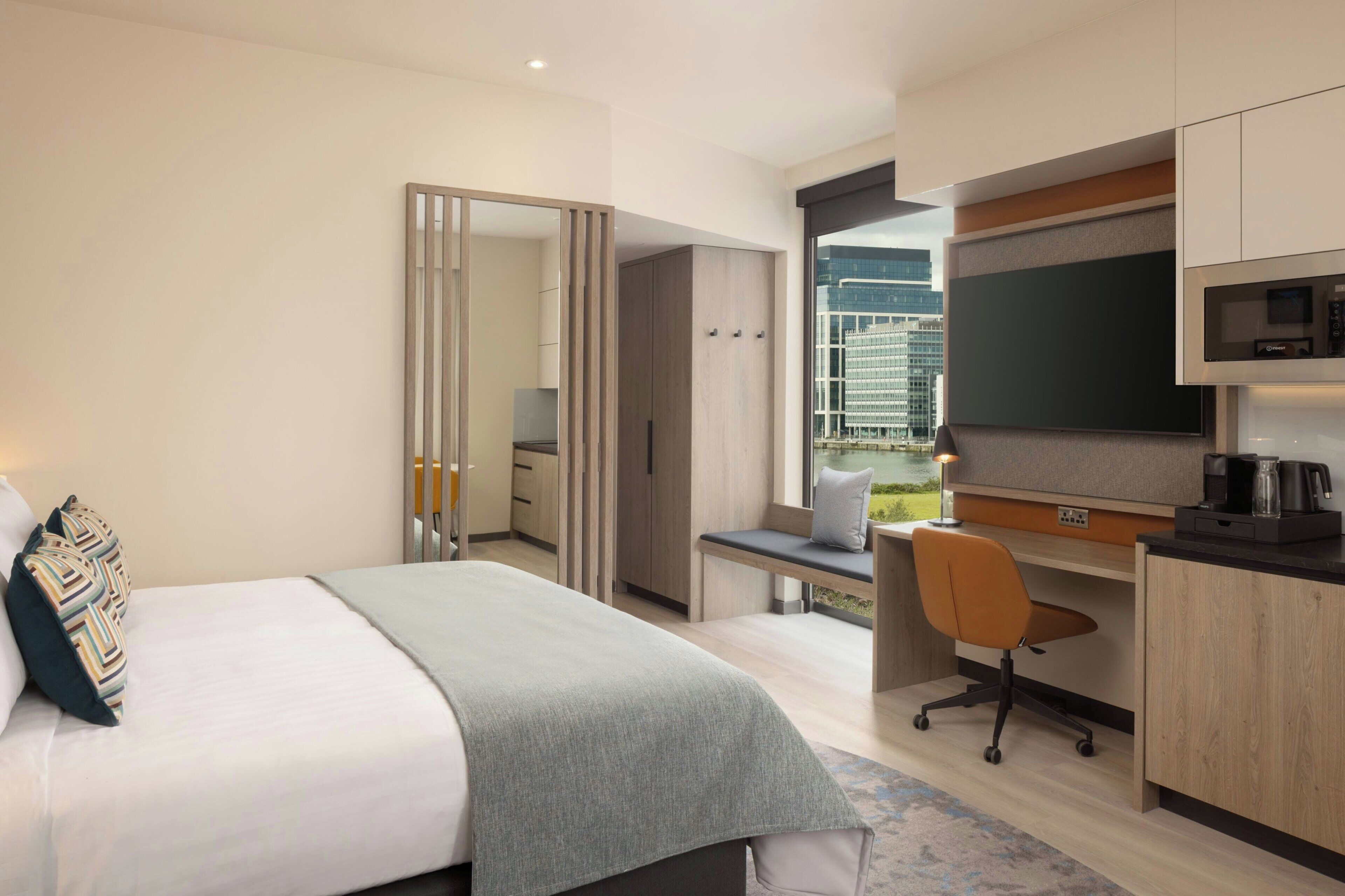 A modern hotel room with a bed, desk, chair, and a large window offering a city view.