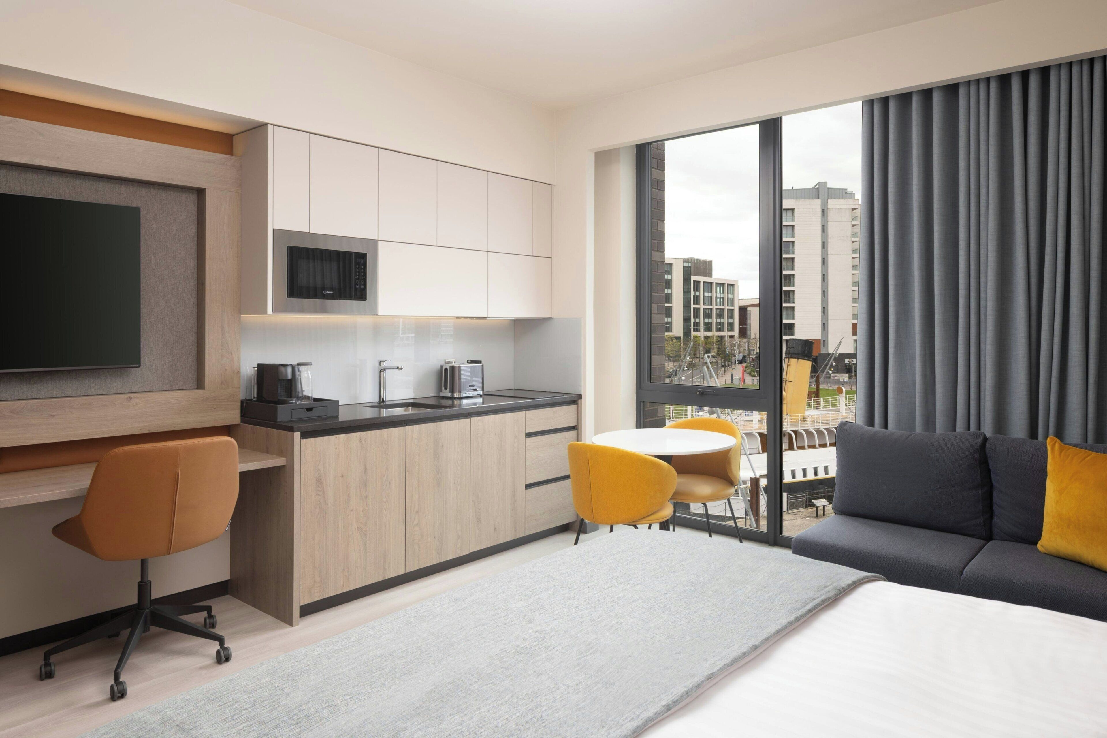 A modern hotel room with a kitchenette, a large bed, a sofa, and a dining area.