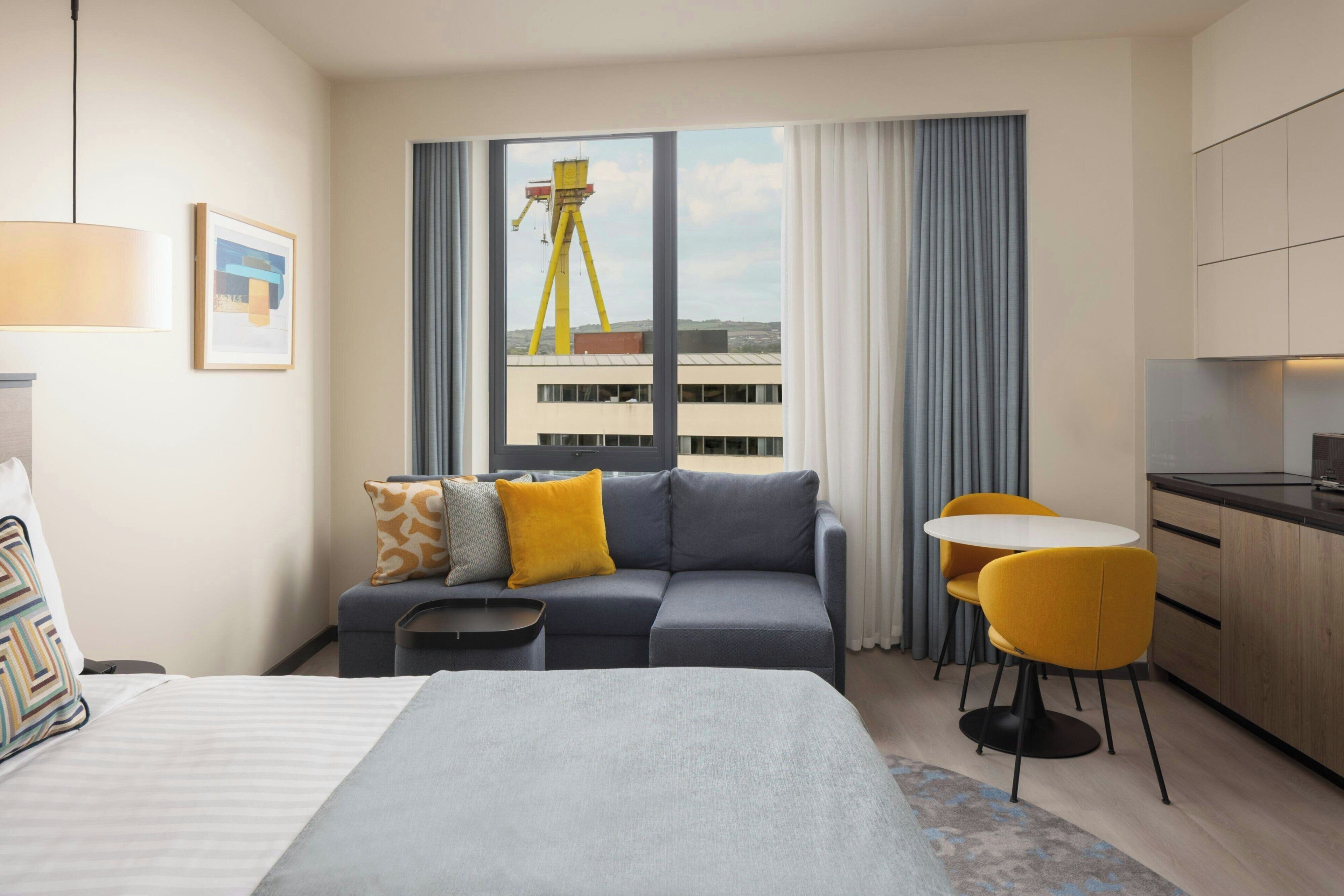 Quarto de hotel com cama, sofá, mesa de jantar com cadeiras, cozinha e uma grande janela com vista para um edifício.
