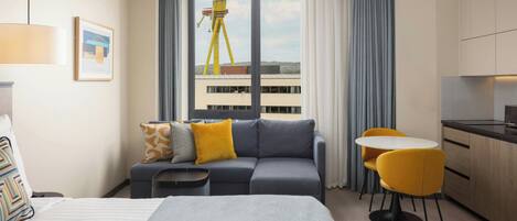 A hotel room with a bed, a sofa, a dining table with chairs, a kitchen area, and a large window with a view of a building.