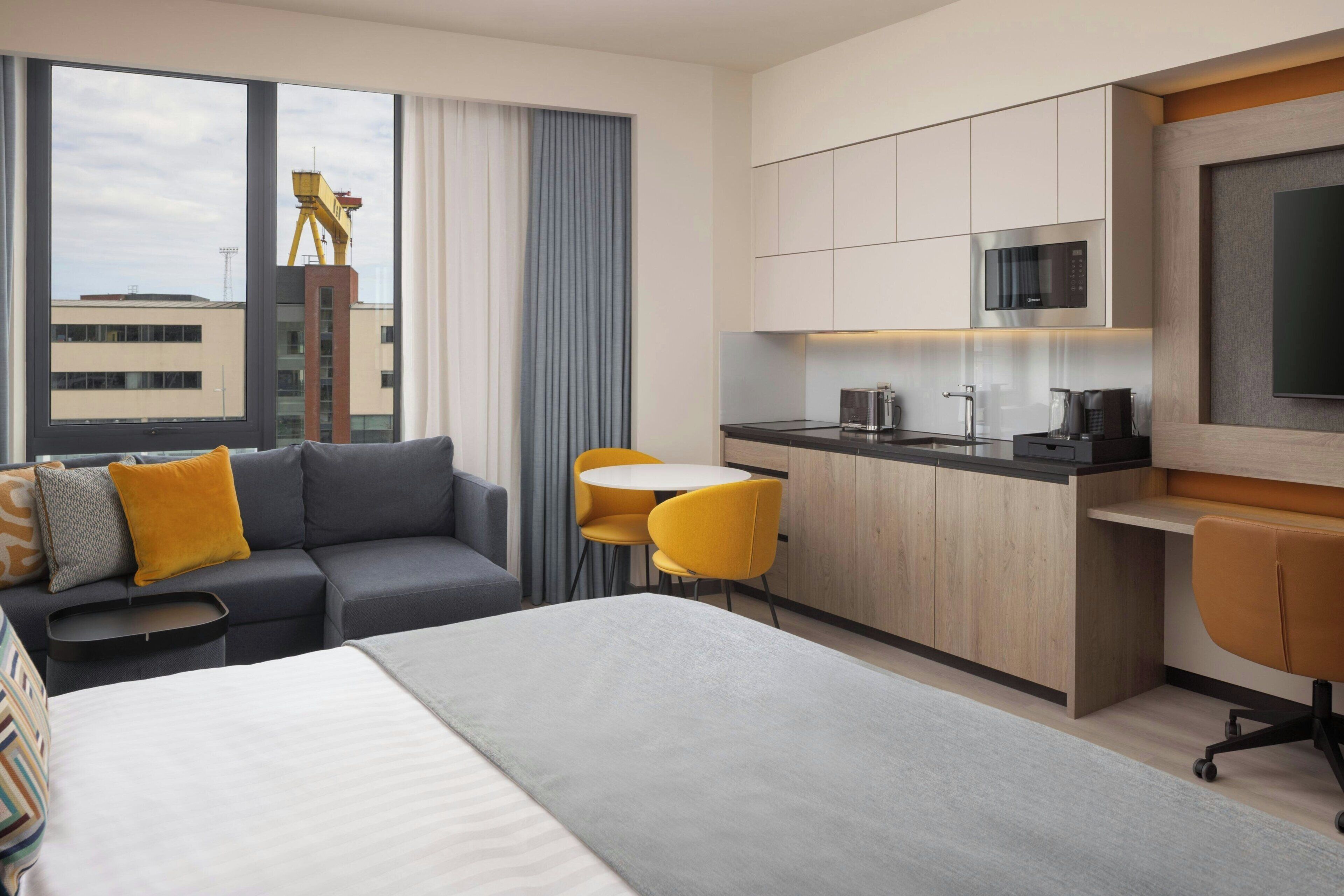 Ein modernes Hotelzimmer mit Bett, Sofa, Esstisch, Stühlen, einer Küchenzeile und einem großen Fenster mit Blick auf Gebäude.