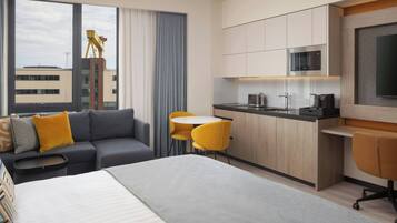 Ein modernes Hotelzimmer mit Bett, Sofa, Esstisch, Stühlen, einer Küchenzeile und einem großen Fenster mit Blick auf Gebäude.
