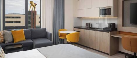 Una habitación de hotel moderna con cama, sofá, mesa de comedor, sillas, kitchenette y un amplio ventanal con vistas a edificios.
