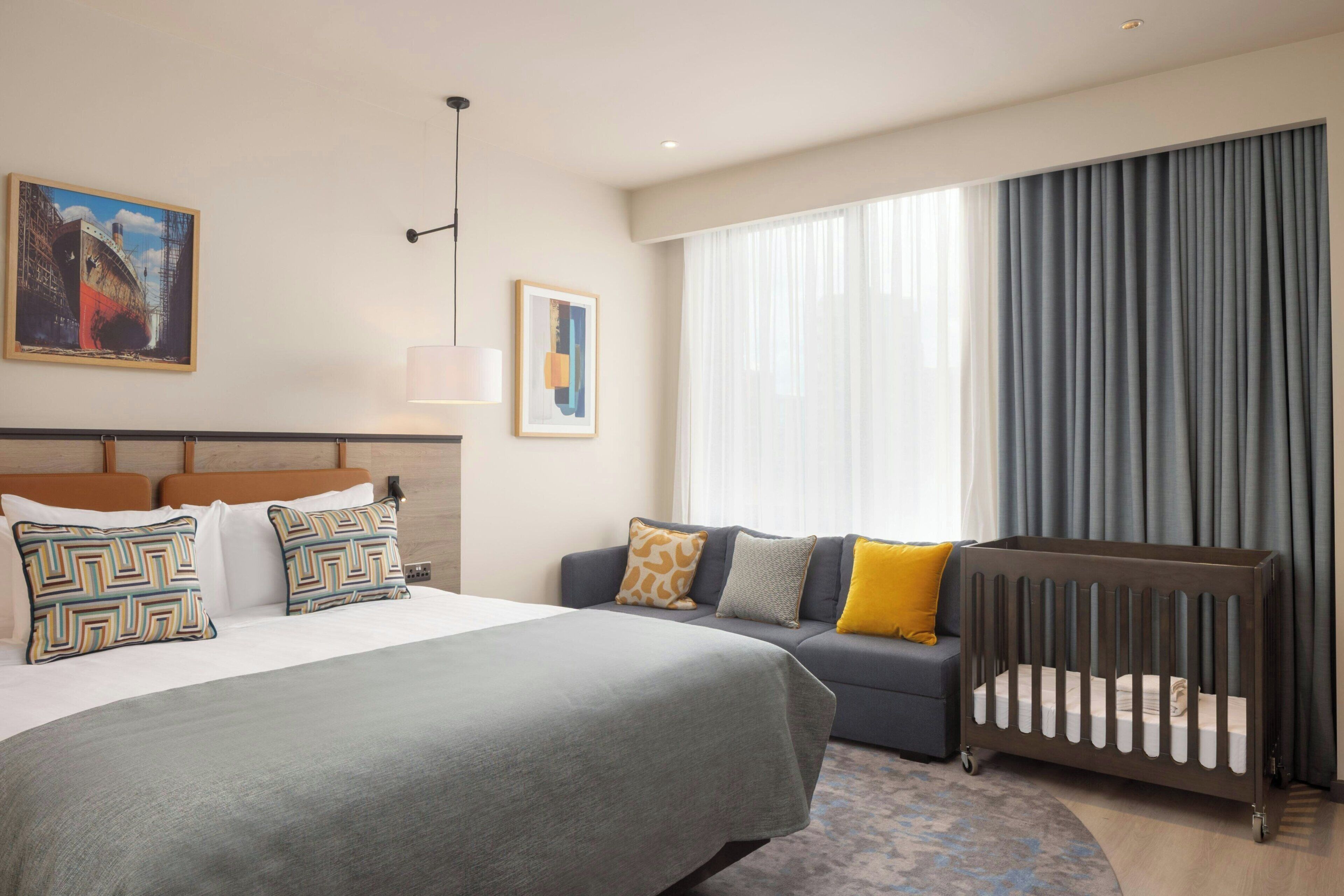 Ein Hotelzimmer mit Bett, einer Schlafcouch, einem Babybett, einem Gemälde und einem Vorhangfenster.