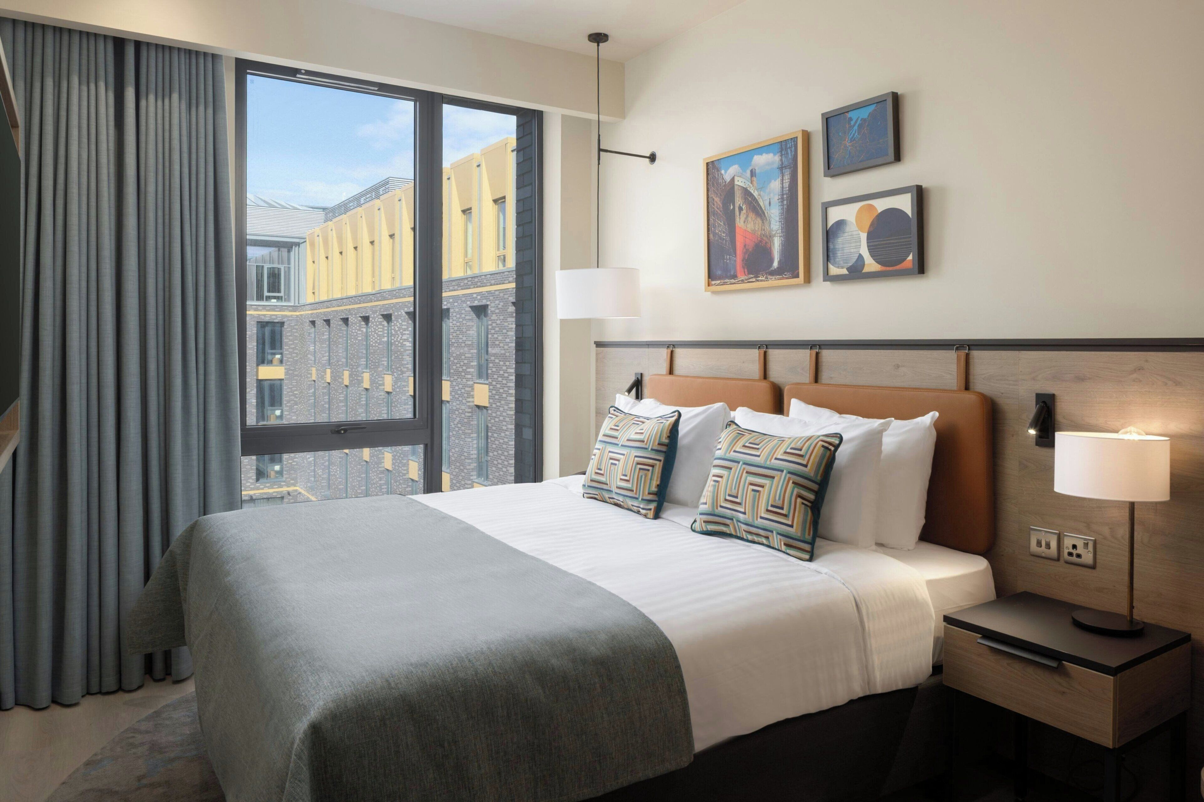 Une chambre d’hôtel avec un grand lit, une table de chevet, une lampe et une vue sur les immeubles par la fenêtre.